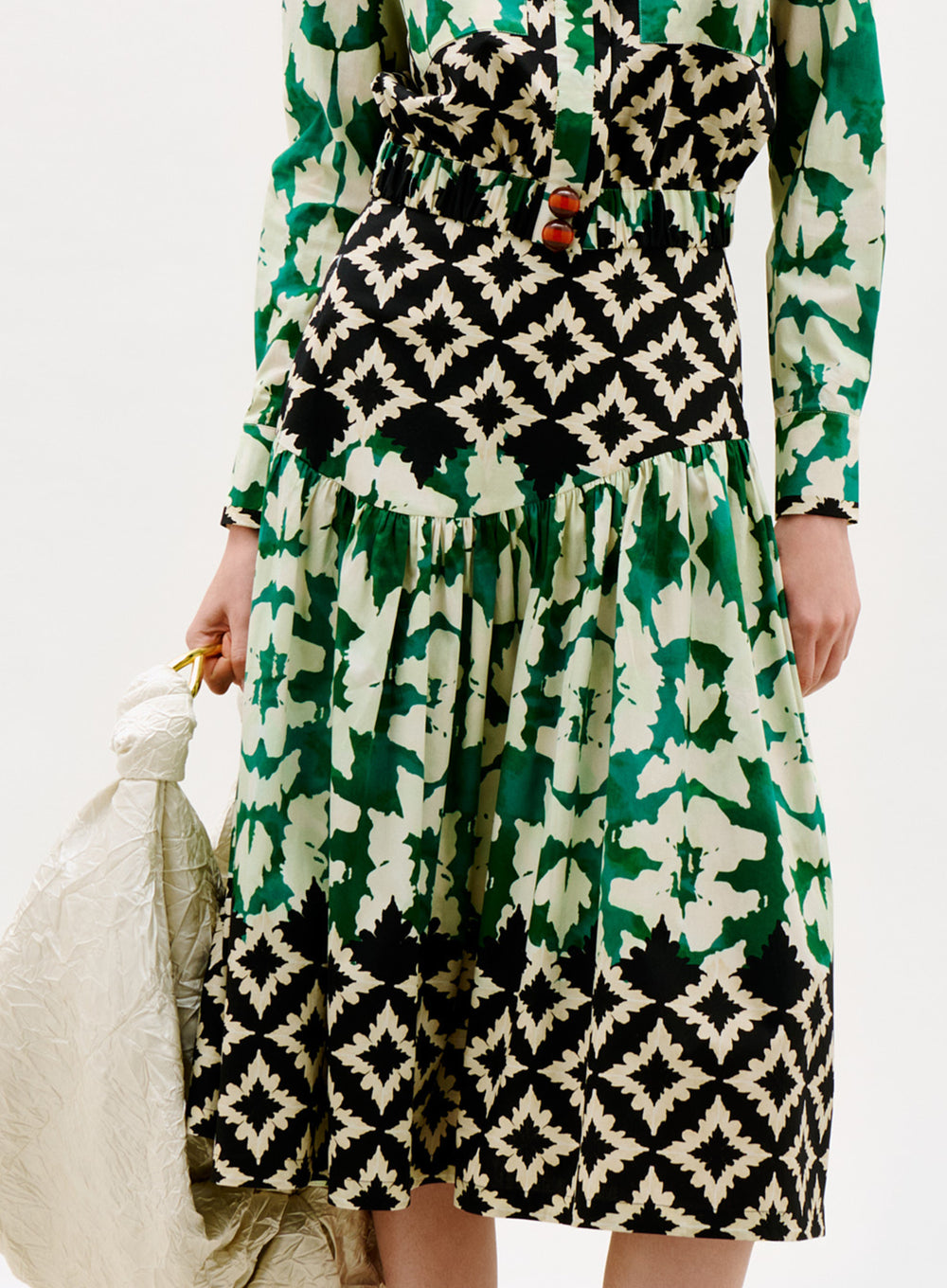 A person wearing the Libby Skirt, featuring green, black, and white geometric and abstract patterns, holds a crinkled light-colored bag at their side.
