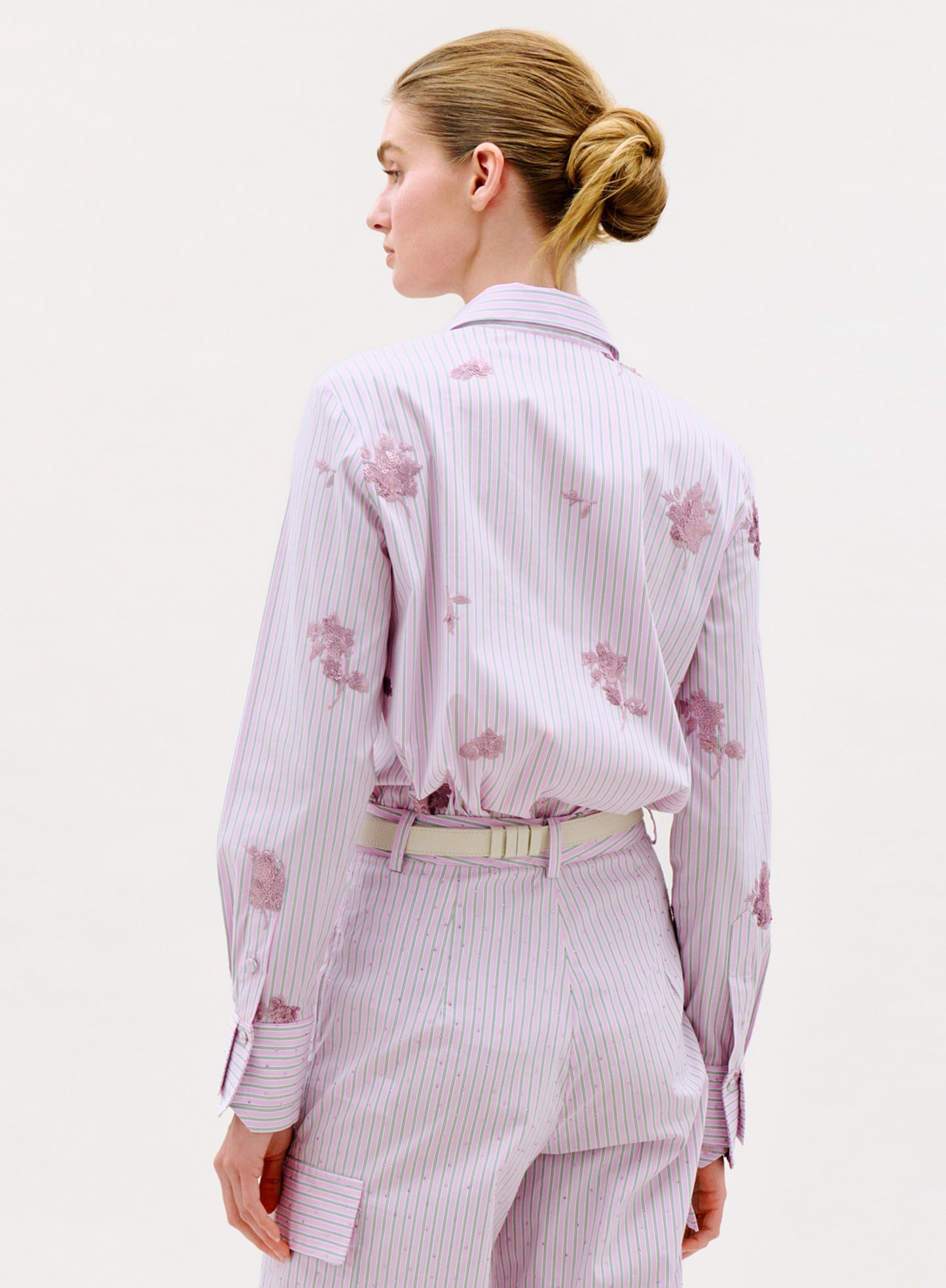 A person with blonde hair in a bun is seen from behind, wearing the Linette Blouse in light purple pinstripes and matching floral pants against a plain, light-colored background.