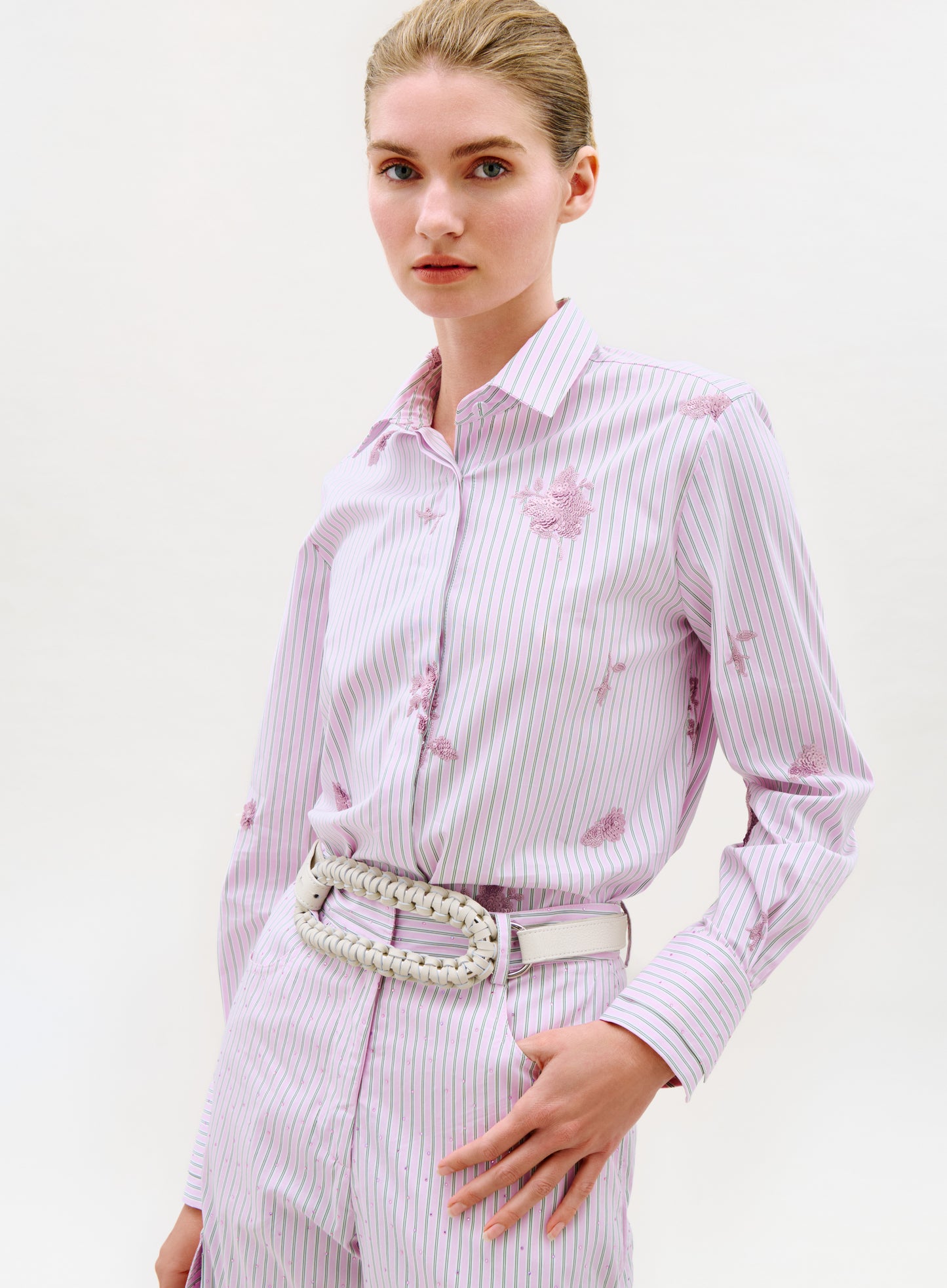 A woman stands against a plain white background wearing the Linette Blouse, a light pink and white striped button-up with subtle floral patterns, paired with matching pants and a white braided belt.
