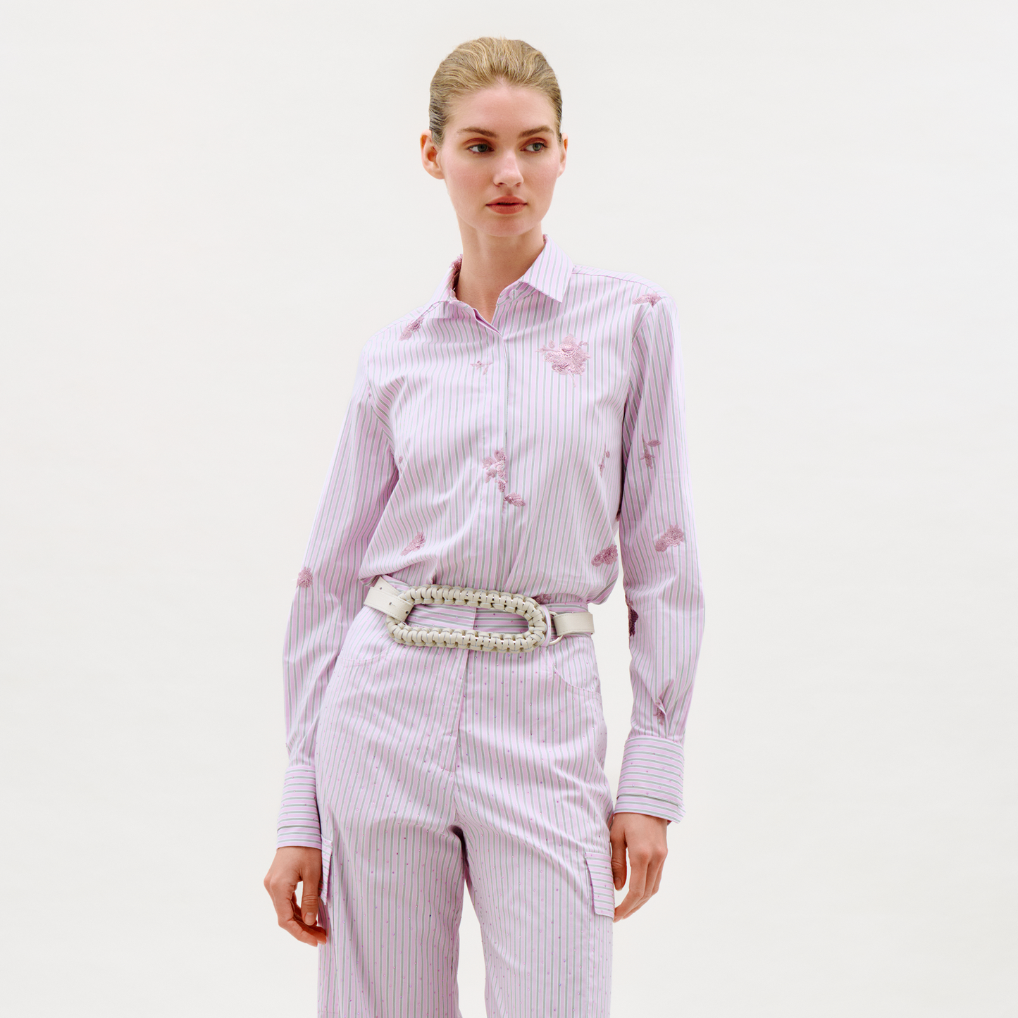A woman stands against a plain background, wearing the Linette Blouse—a light pink and white striped button-down shirt with floral patterns—along with matching striped pants and a wide braided belt. Her hair is pulled back as she gazes slightly to the side.