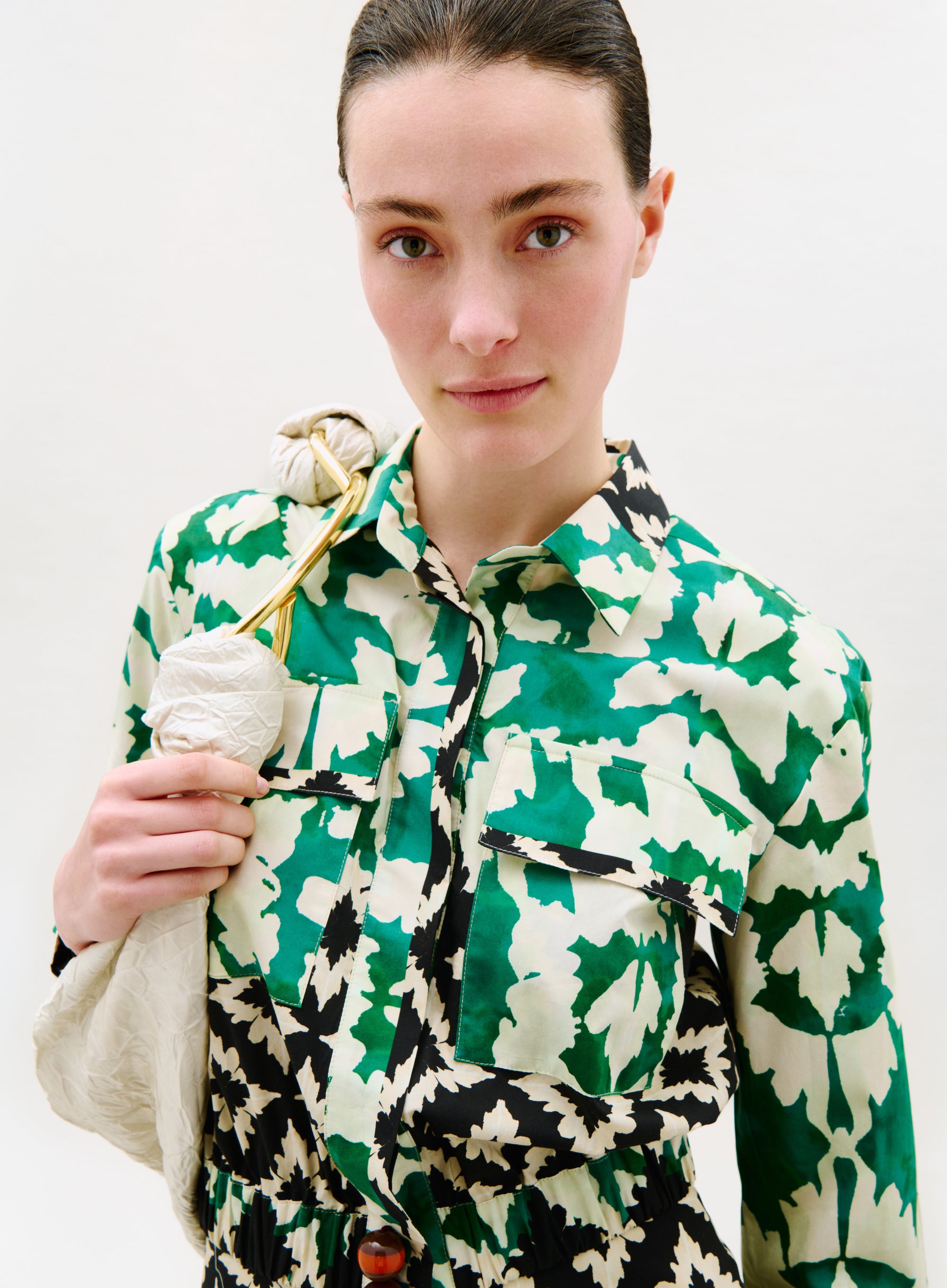 A woman with fair skin and dark hair pulled back models the Soleil Blouse, featuring a green, white, and black pattern. She holds a white bag with a gold strap over her shoulder, standing against a plain light background.