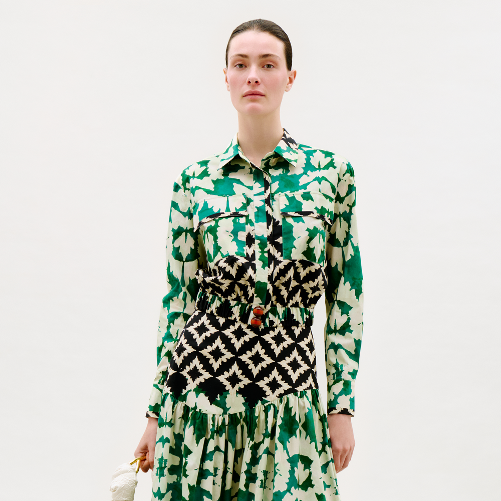 A person stands against a plain background, wearing the Soleil Blouse—a long-sleeved top with green and white leafy patterns combined with black and white geometric designs—holding a small white object in their left hand.