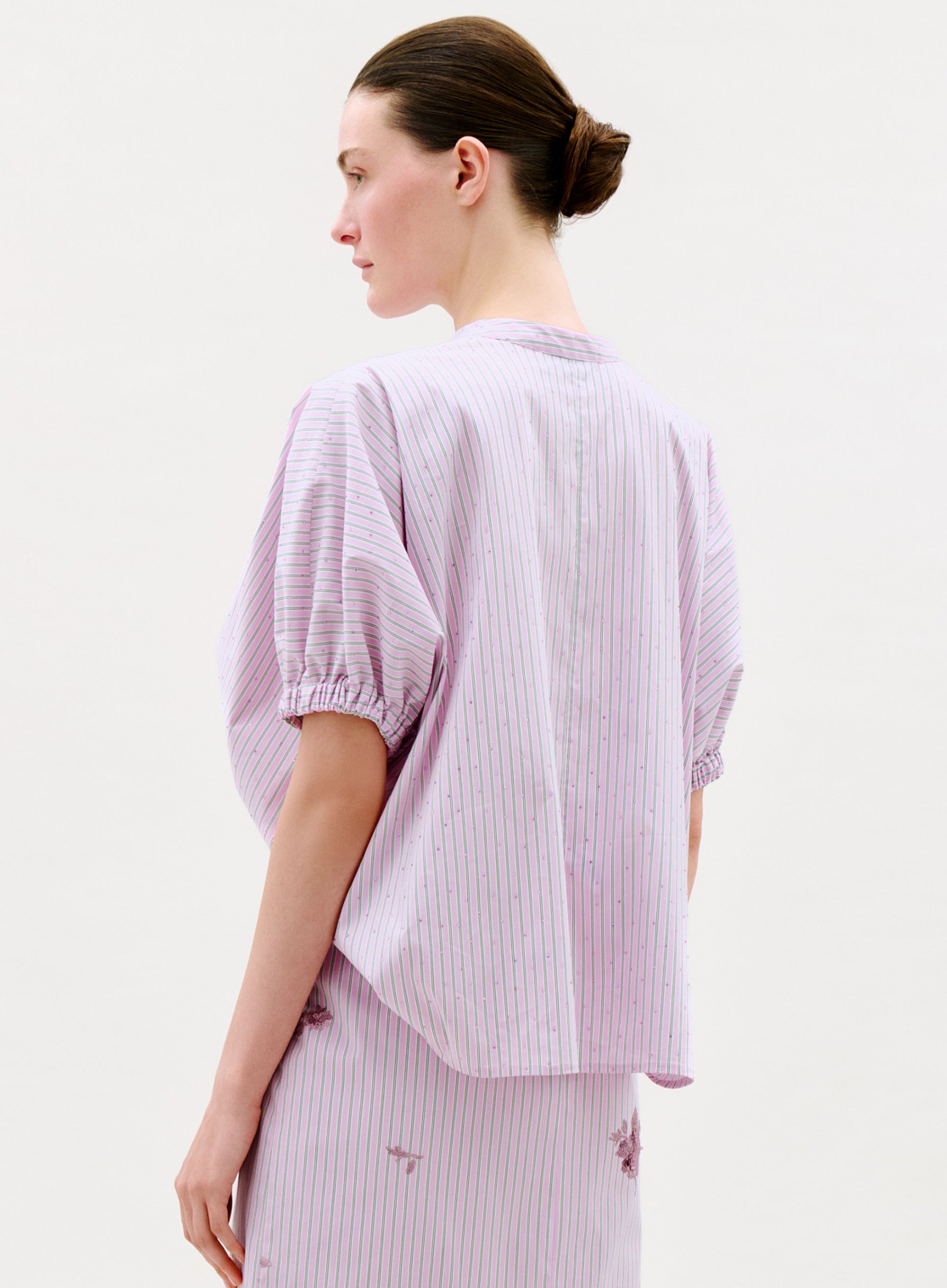 A woman with brown hair in a low bun wears the loose-fitting Susane Blouse, featuring short sleeves and light purple stripes, paired with a matching skirt. She stands sideways against a plain background, looking to the left.