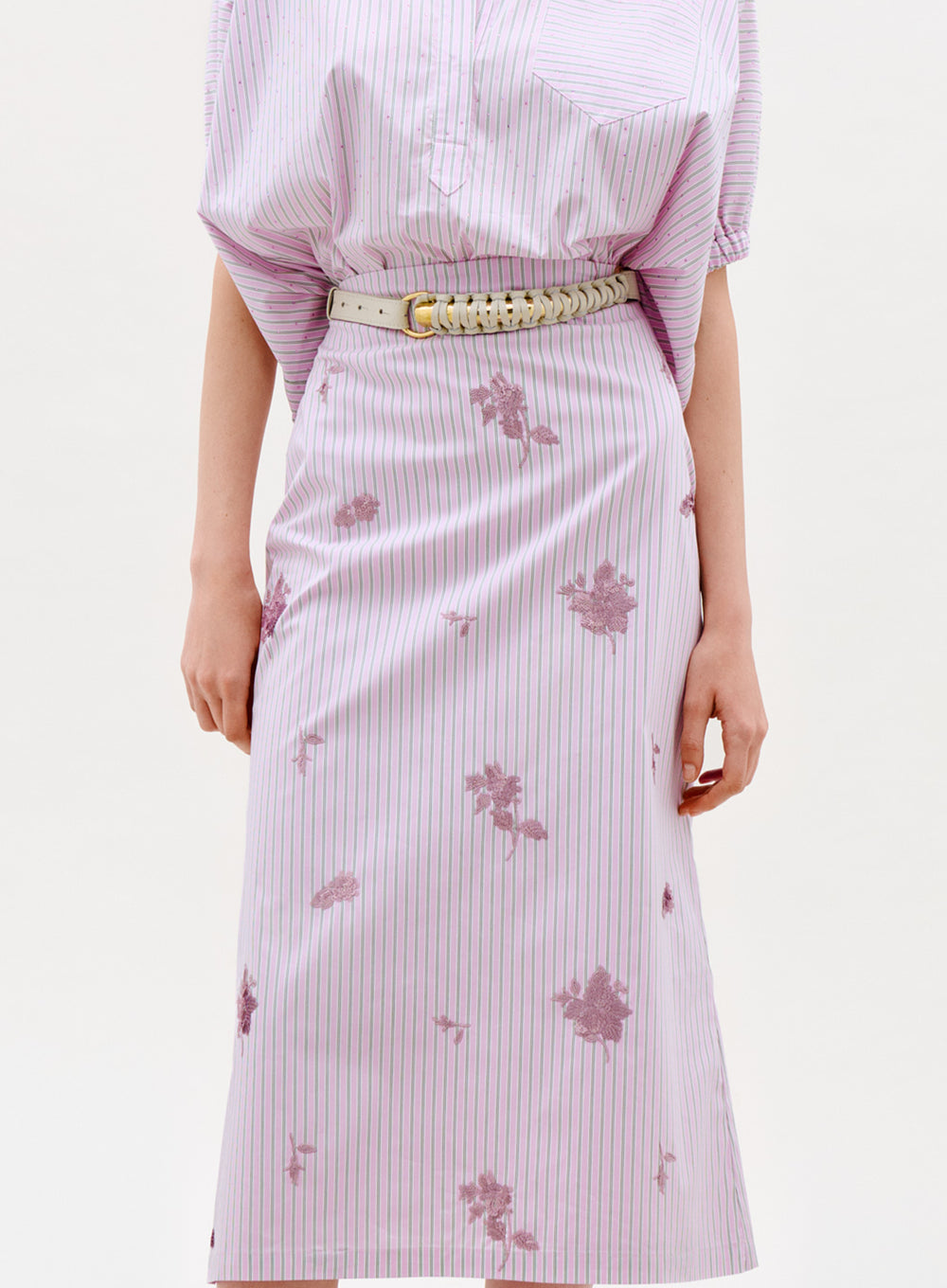 Wearing the Atira Skirt in light purple and white stripes with floral patterns, paired with a matching top and beige woven belt. The photo shows a shoulder-to-knee view against a plain background.