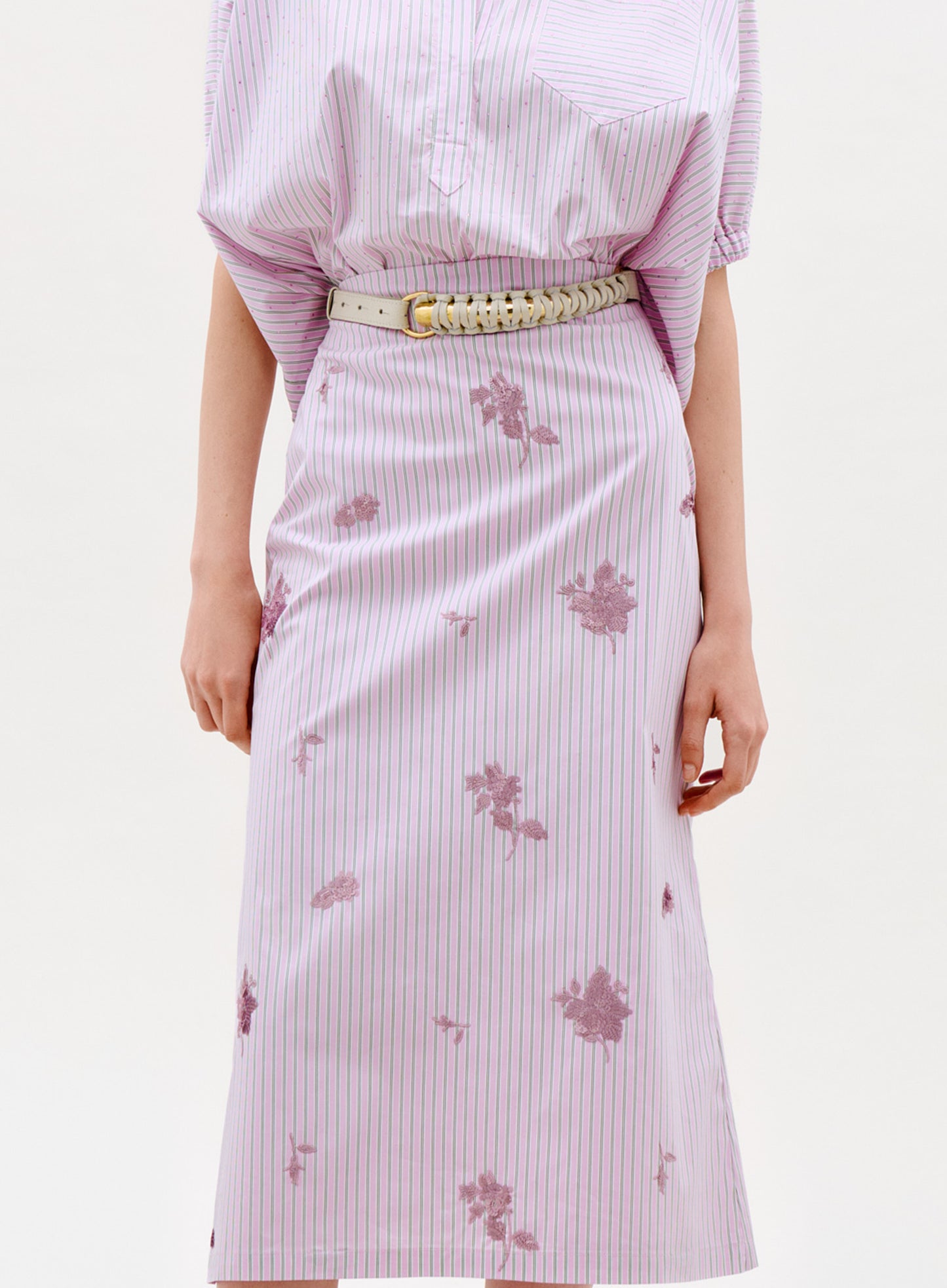 Wearing the Atira Skirt in light purple and white stripes with floral patterns, paired with a matching top and beige woven belt. The photo shows a shoulder-to-knee view against a plain background.