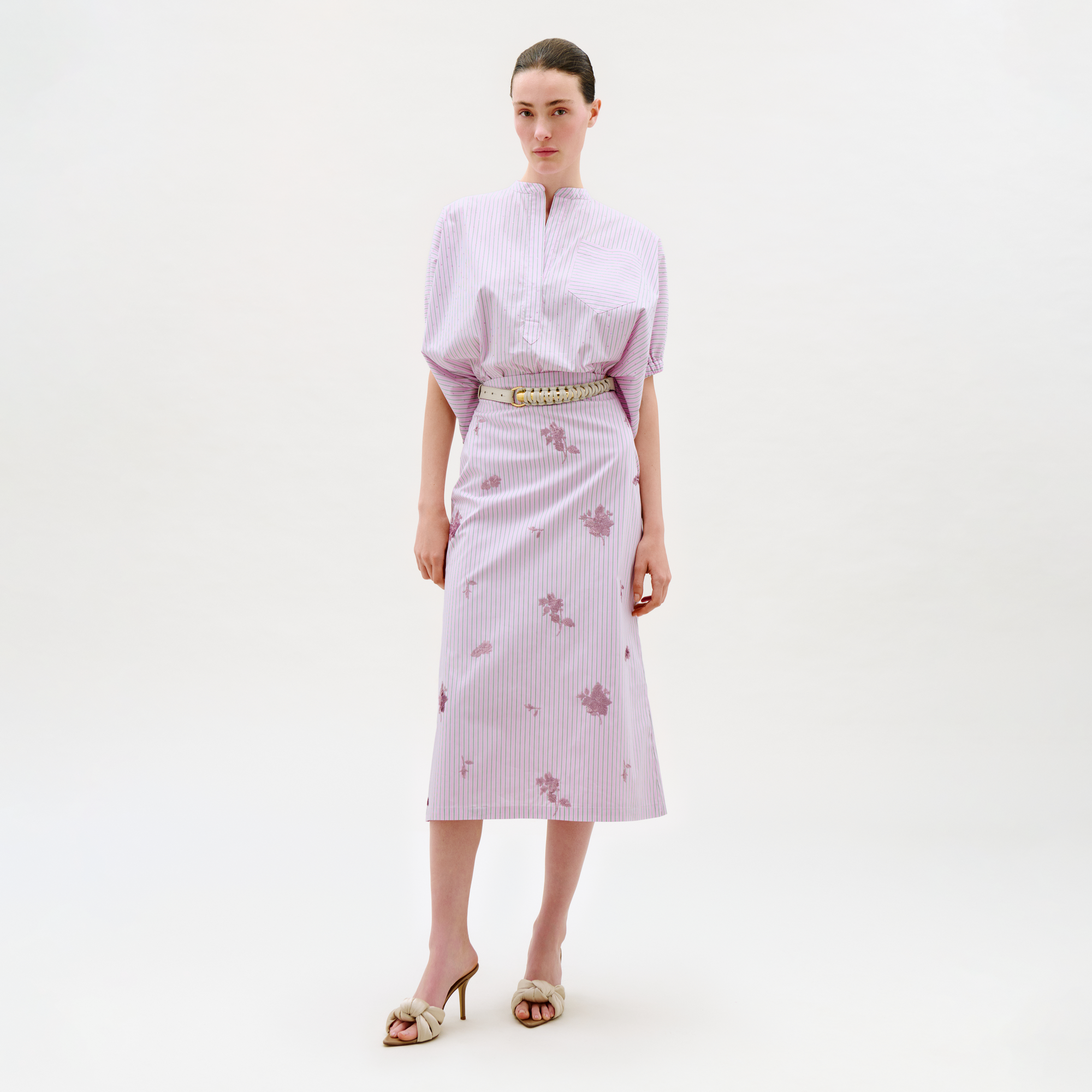 A woman wears the Atira Skirt, a light purple striped midi skirt with subtle floral prints, paired with a matching puff-sleeve blouse and heeled sandals. She stands against a plain white background with a neutral expression.
