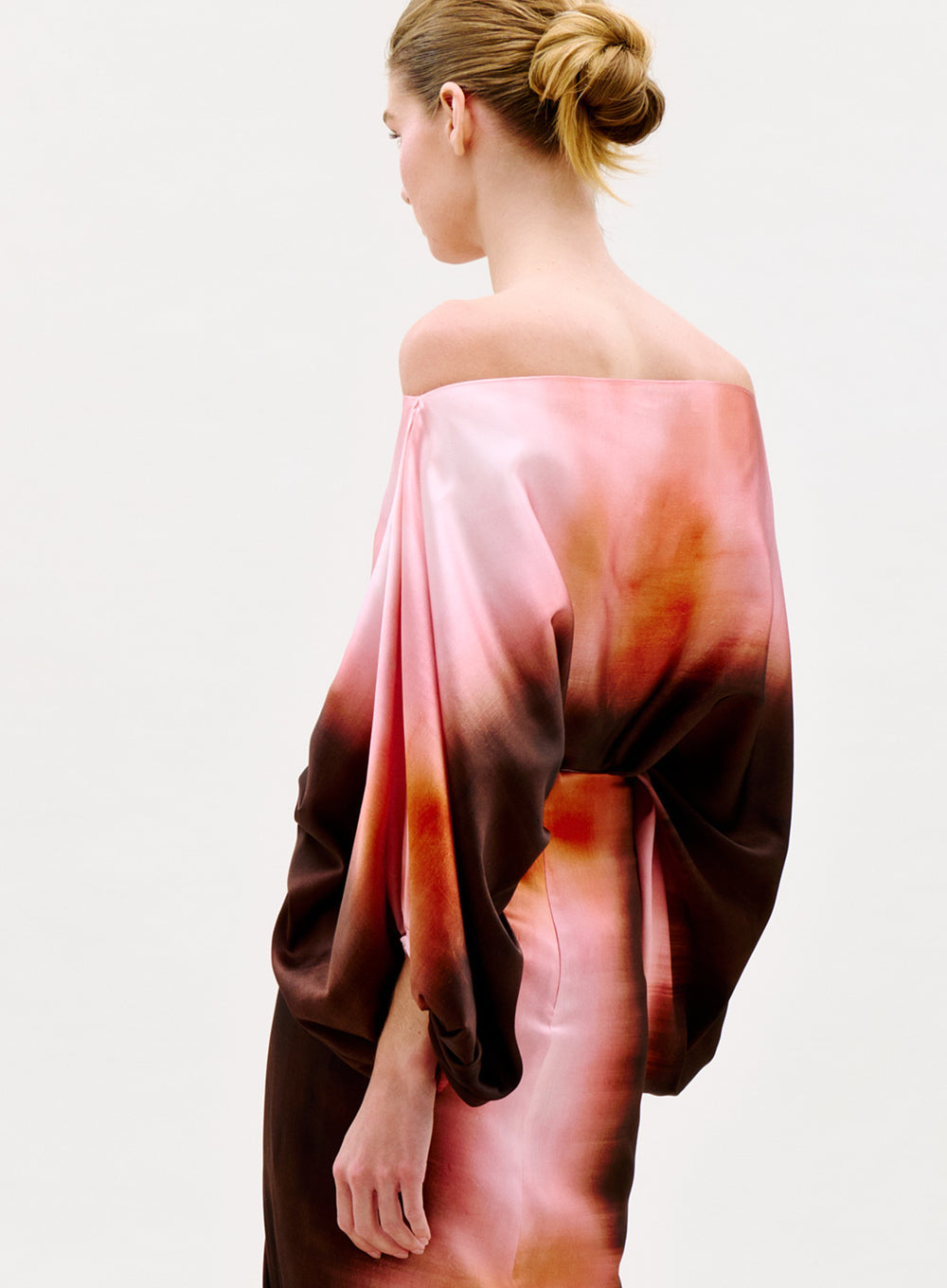A woman with her hair in a bun wears the Bellagio Blouse, an off-the-shoulder top with wide sleeves and a pink, orange, and brown gradient pattern, standing against a plain light background.