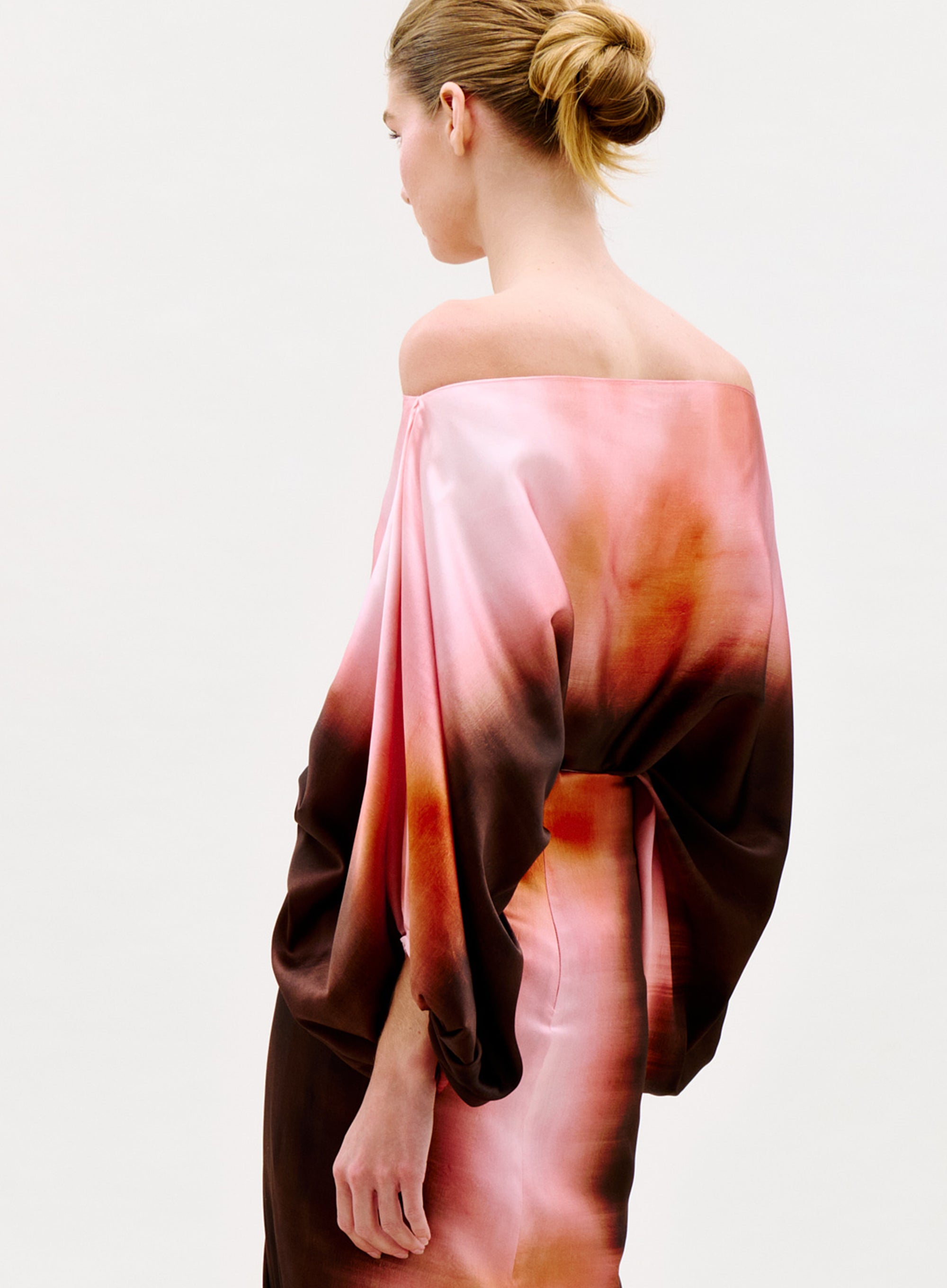 A woman with her hair in a bun wears the Bellagio Blouse, an off-the-shoulder top with wide sleeves and a pink, orange, and brown gradient pattern, standing against a plain light background.