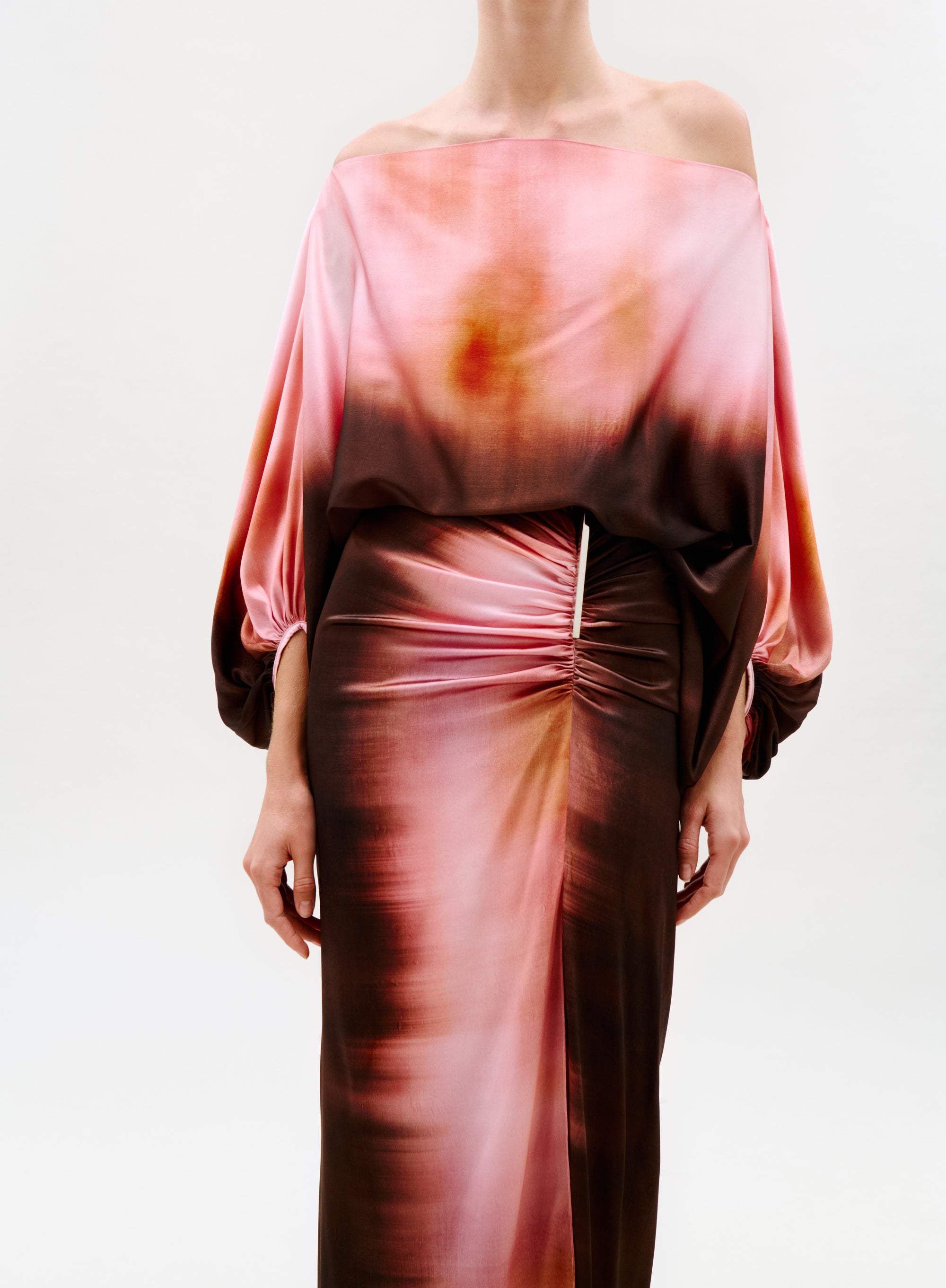 A person wearing the Bellagio Blouse, an off-the-shoulder, long-sleeve top with a pink, brown, and orange gradient pattern, gathered detail, and decorative waist clasp. The background is plain white.