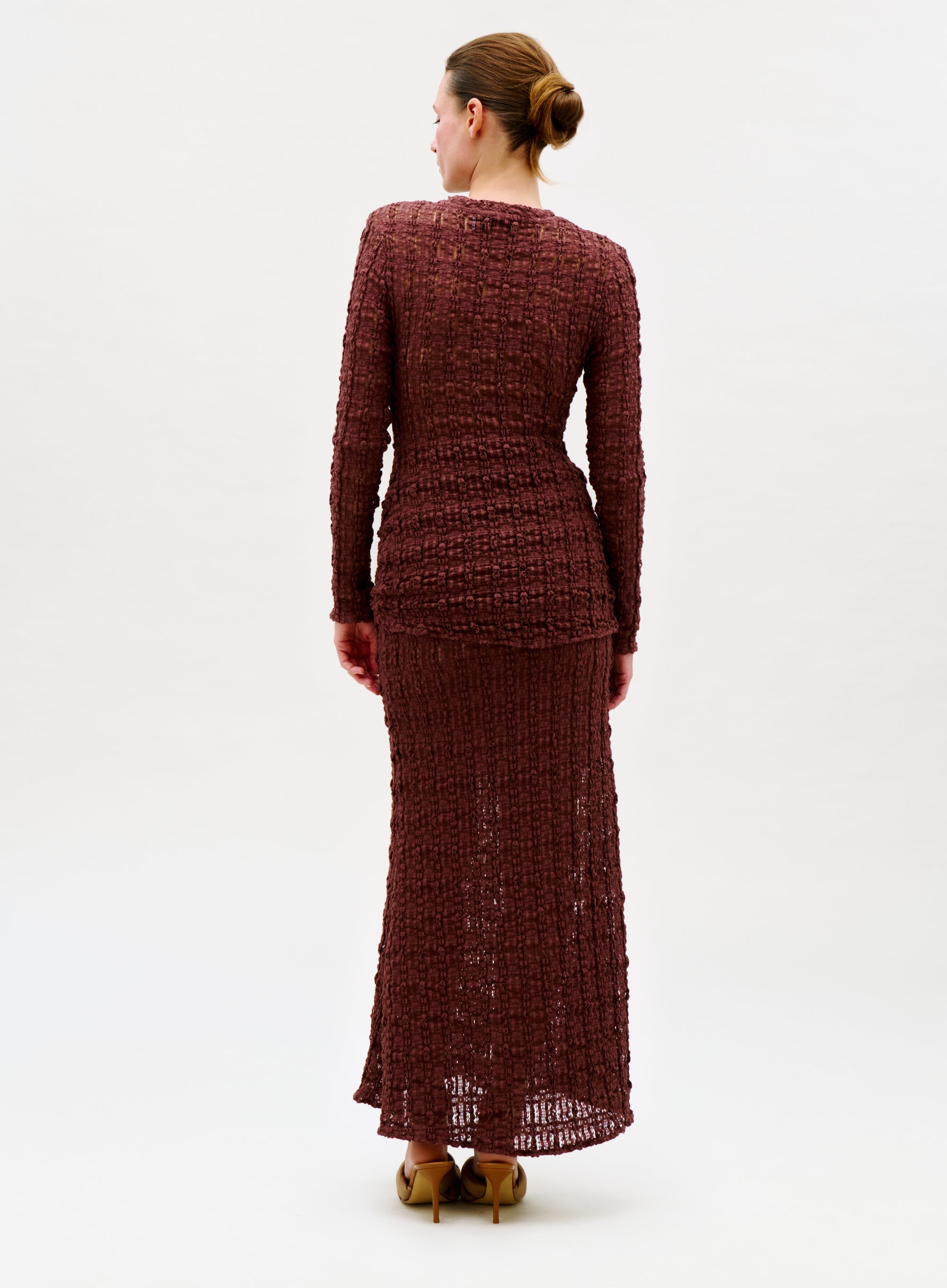 A woman faces away, modeling the Ciji Skirt—a long, textured maroon skirt with a sleek silhouette. She wears tan high heels and has her hair neatly tied back against a plain white background.