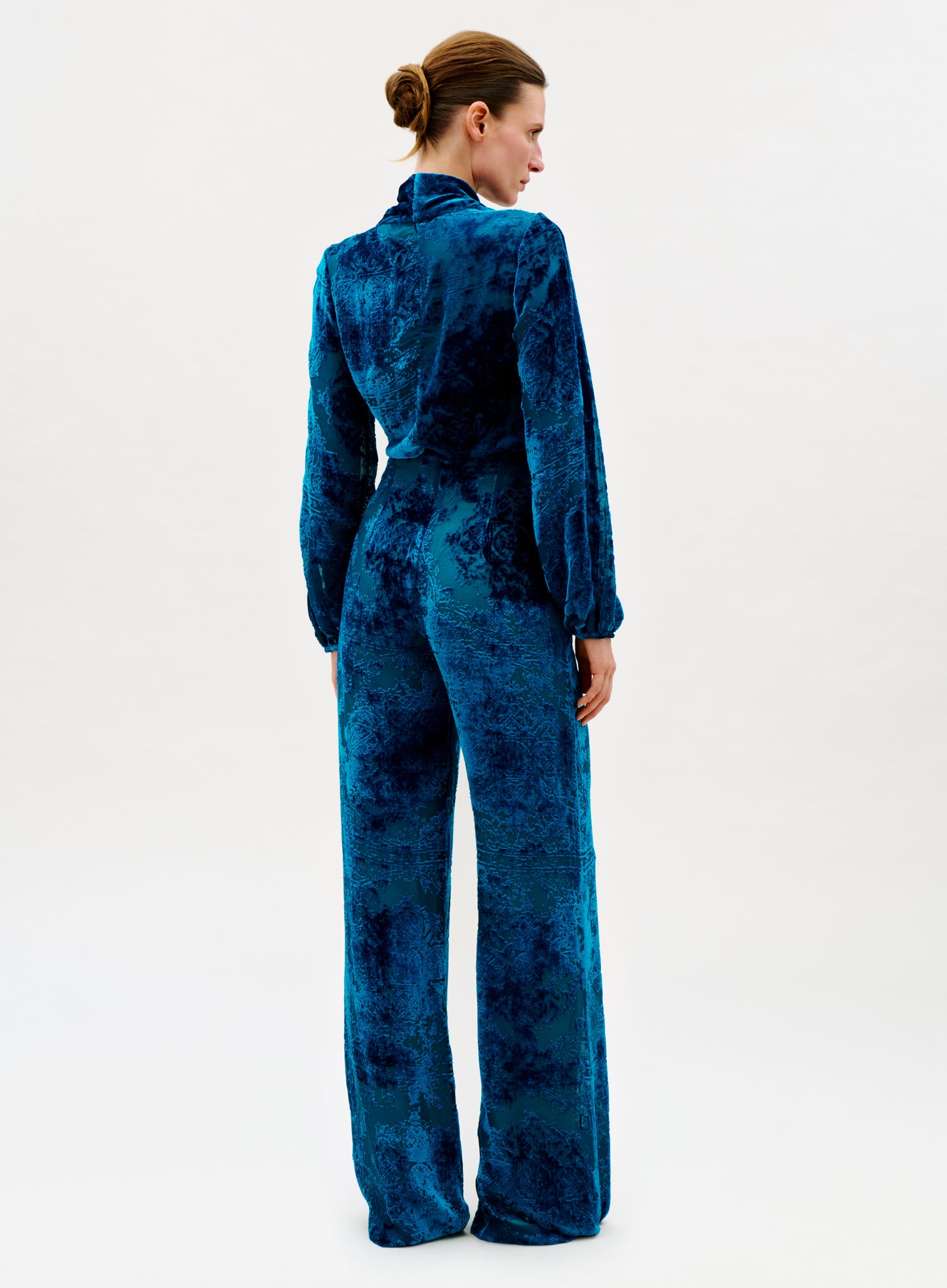 A woman faces away, wearing the high-waisted Como Pant in blue velvet with wide legs. Her hair is tied back in a neat bun as she poses against a plain, light background.