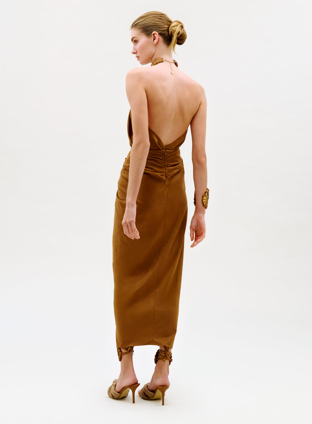 A woman stands facing away, wearing the Denise Skirt in brown with gathered details and high heels. Her hair is styled in a bun, and she accessorizes with a chunky bracelet and necklace against a plain, light background.