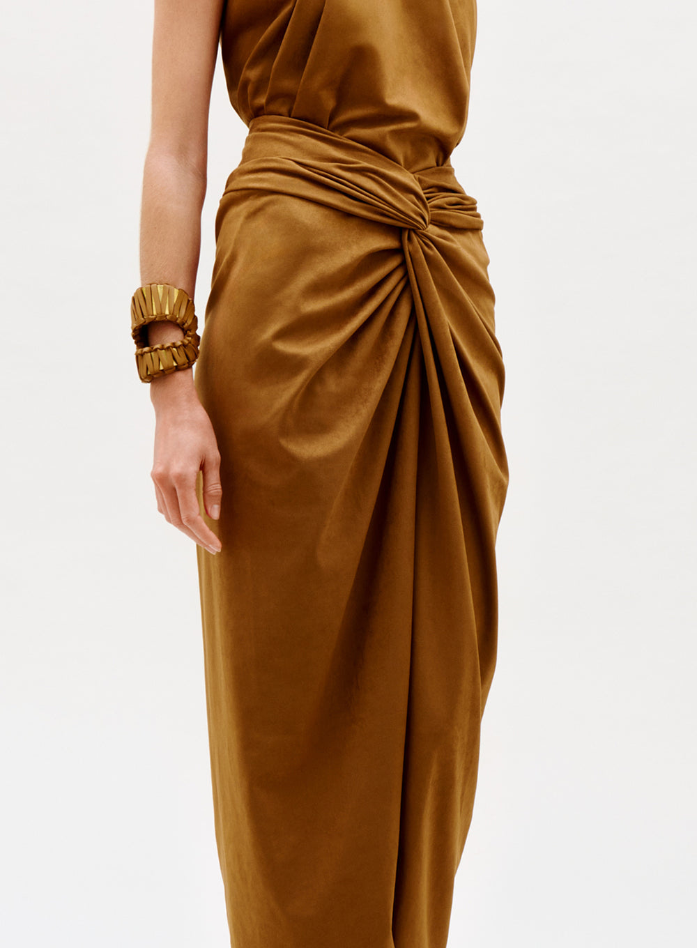 A person models the Denise Skirt, a golden-brown draped piece paired with a matching sleeveless top and chunky gold bracelet, standing against a plain white background.