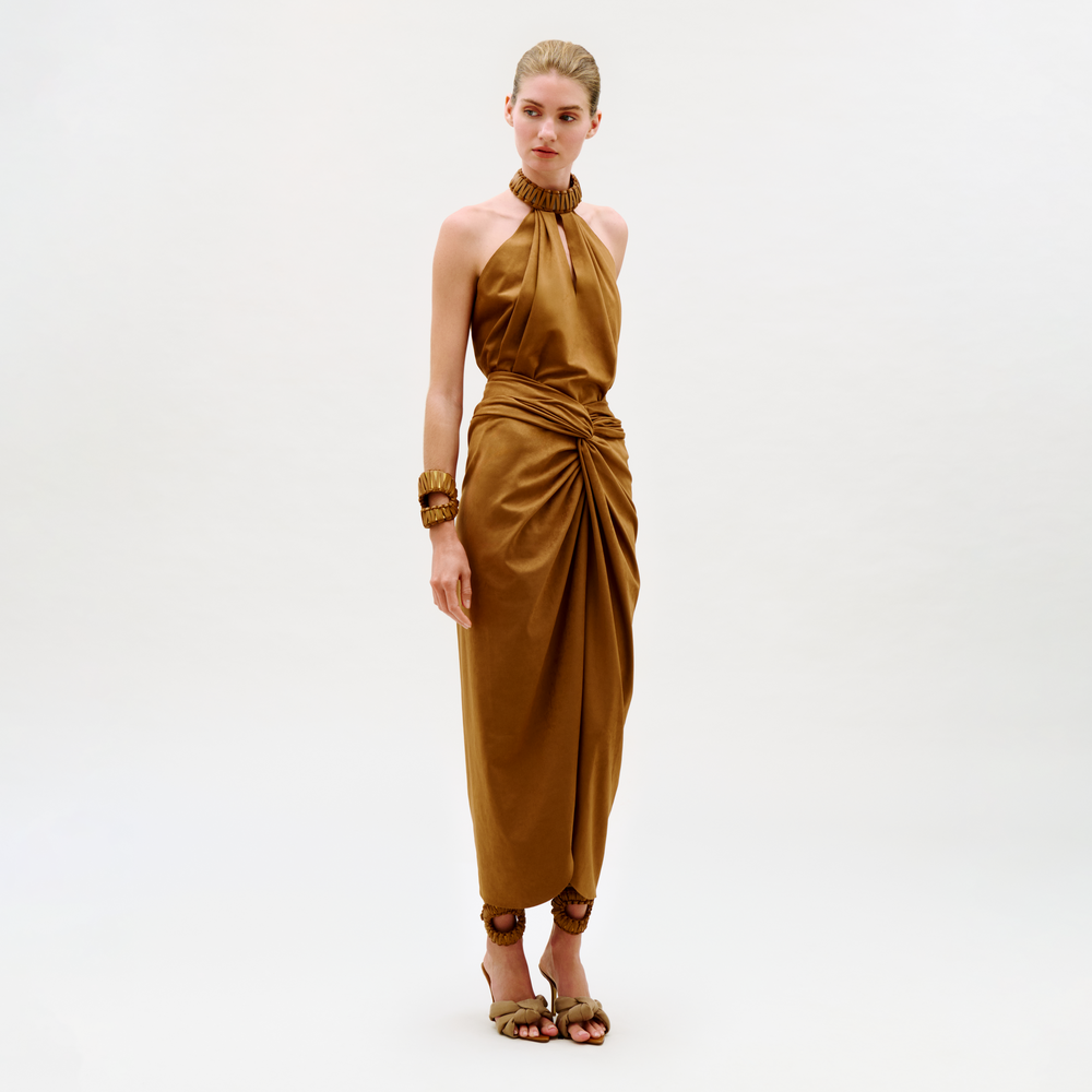 A woman stands against a plain background wearing the Denise Skirt, a golden-brown halter style with a gathered waist and draped skirt. She completes her look with matching bracelets and heeled sandals, gazing softly ahead.