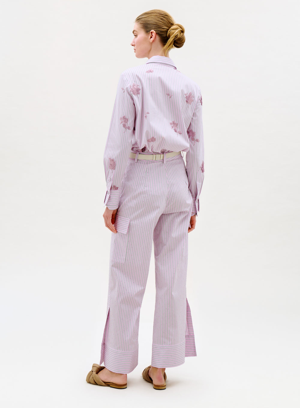 A woman with her hair in a bun wears the Falak Pant—pale pink and white stripes with floral patterns, wide legs, and slits at the hem—along with a matching top and tan slip-on shoes. She stands facing away.