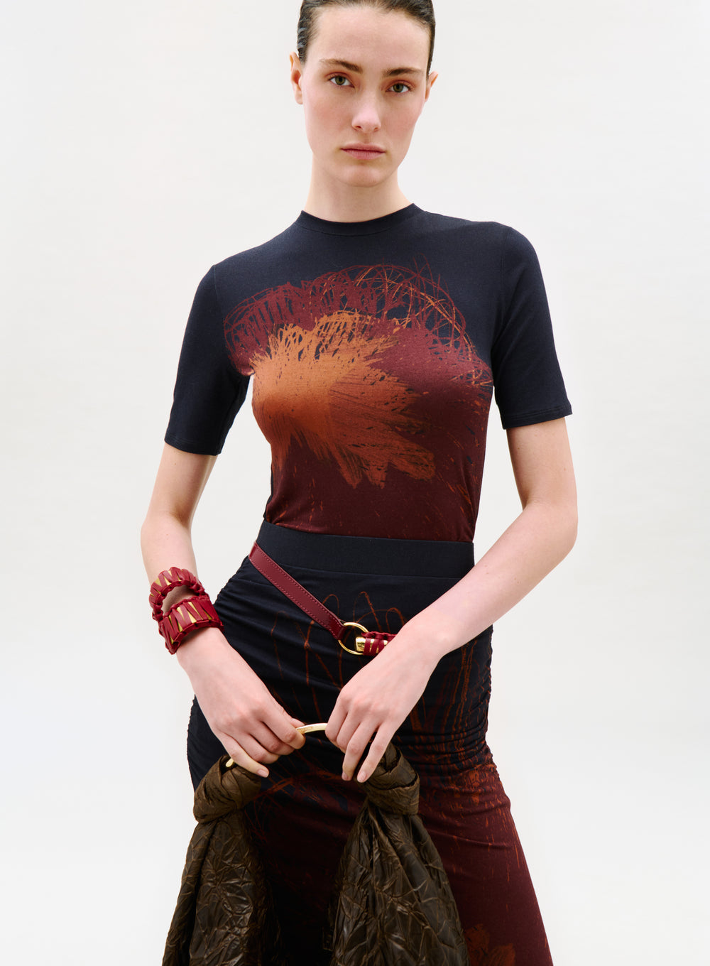 A woman with dark hair pulled back wears the Fano T-Shirt with an abstract red and orange design. She holds a brown textured jacket, carries a red bag, and poses against a plain white background.