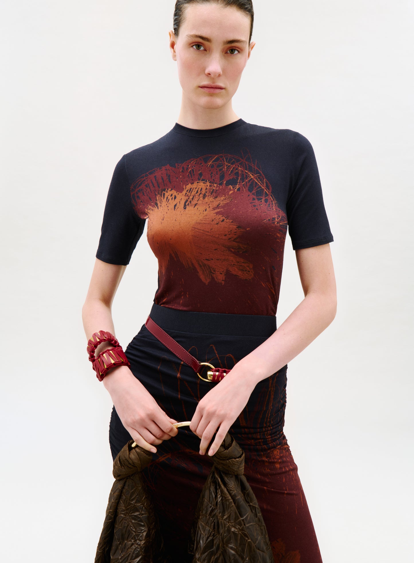 A woman with dark hair pulled back wears the Fano T-Shirt with an abstract red and orange design. She holds a brown textured jacket, carries a red bag, and poses against a plain white background.