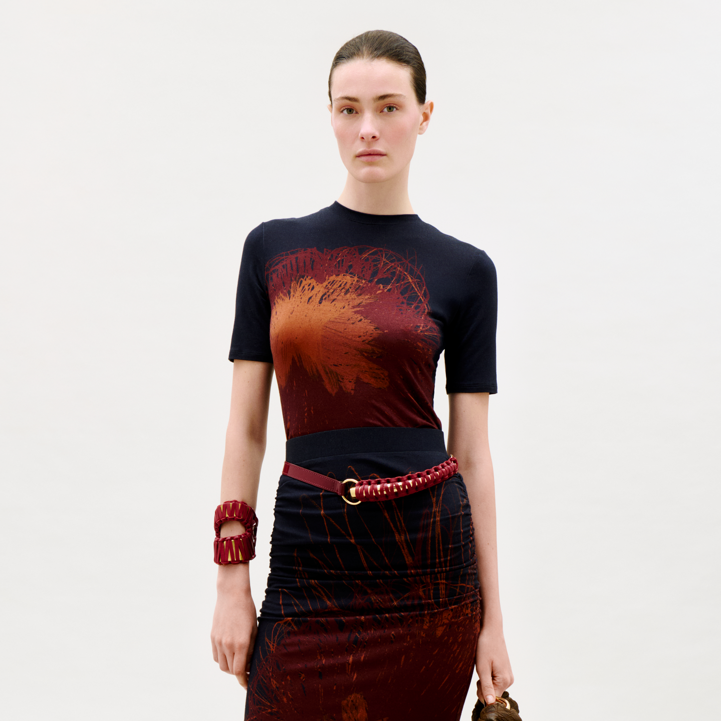 A woman stands against a plain light background, wearing the Fano T-Shirt, paired with a red braided belt and chunky red bracelet, holding a handbag and looking directly at the camera.