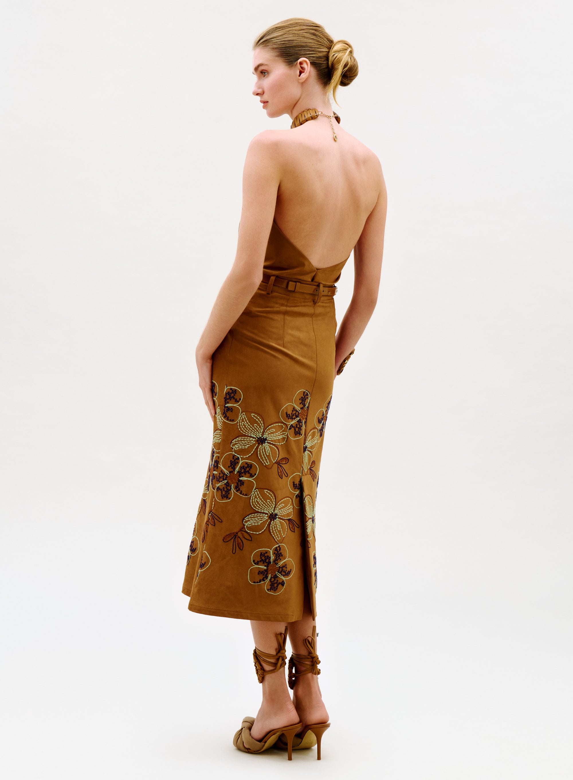 A woman stands in profile, wearing the Layan Skirt with a tan, floral design. She pairs it with matching high-heeled sandals and wears her hair in a neat bun, posing against a simple light background.