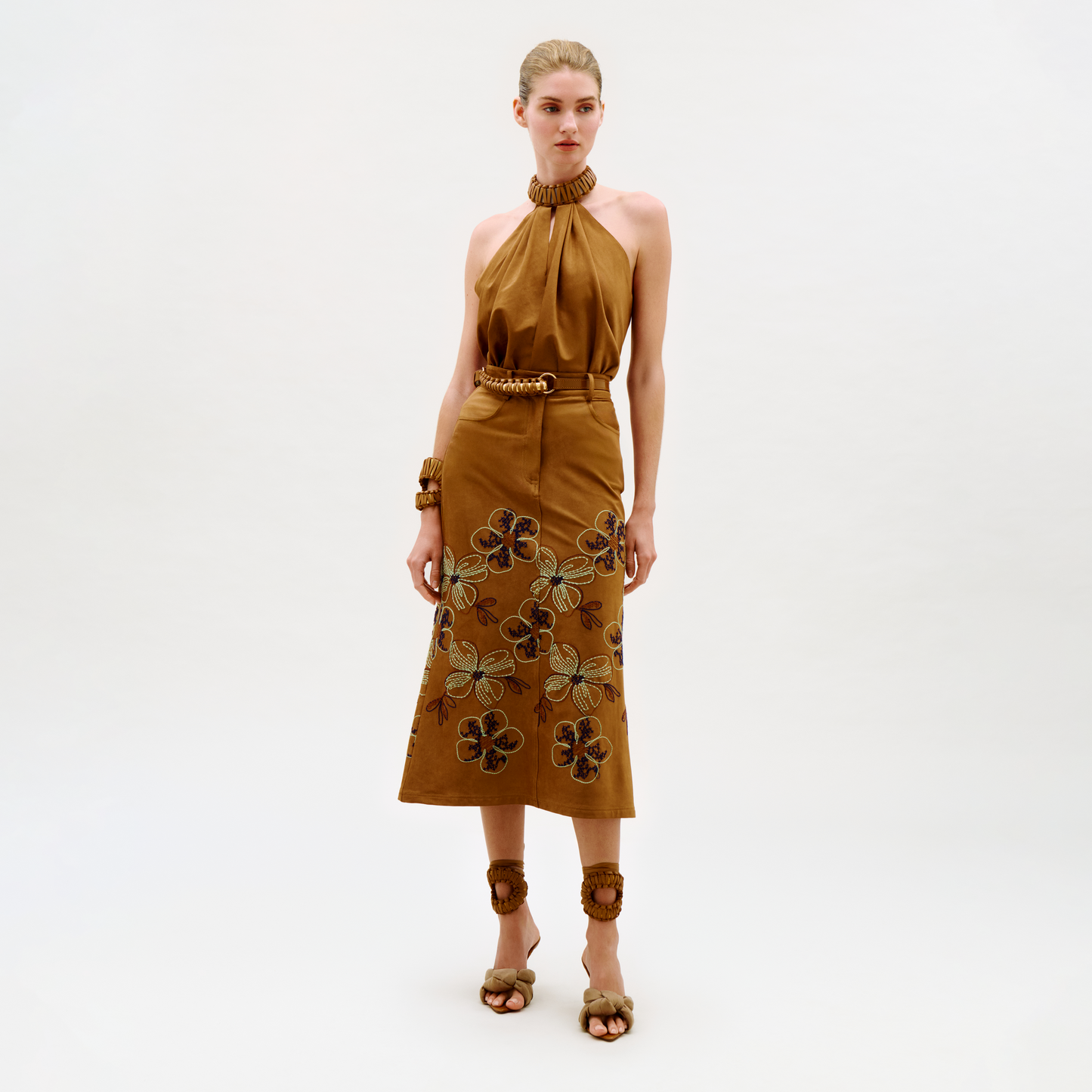 A woman stands against a plain background wearing the Layan Skirt, styled with brown sandals featuring floral accents, a matching belt, and bracelets. Her hair is pulled back in a sleek style.