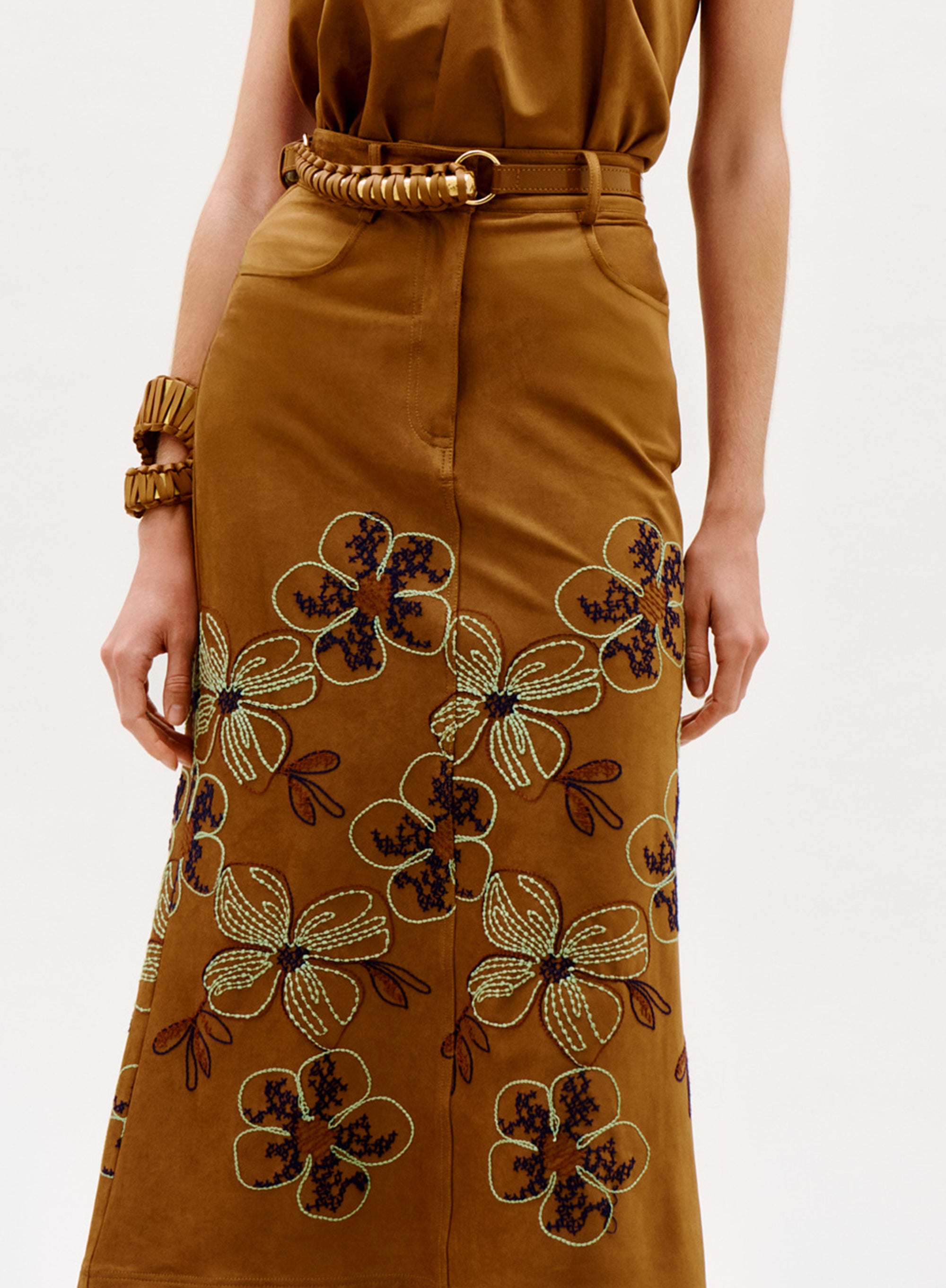 Wearing the Layan Skirt, a brown skirt with large embroidered floral patterns, paired with a matching brown top and accessorized with a chunky brown belt and bracelet.