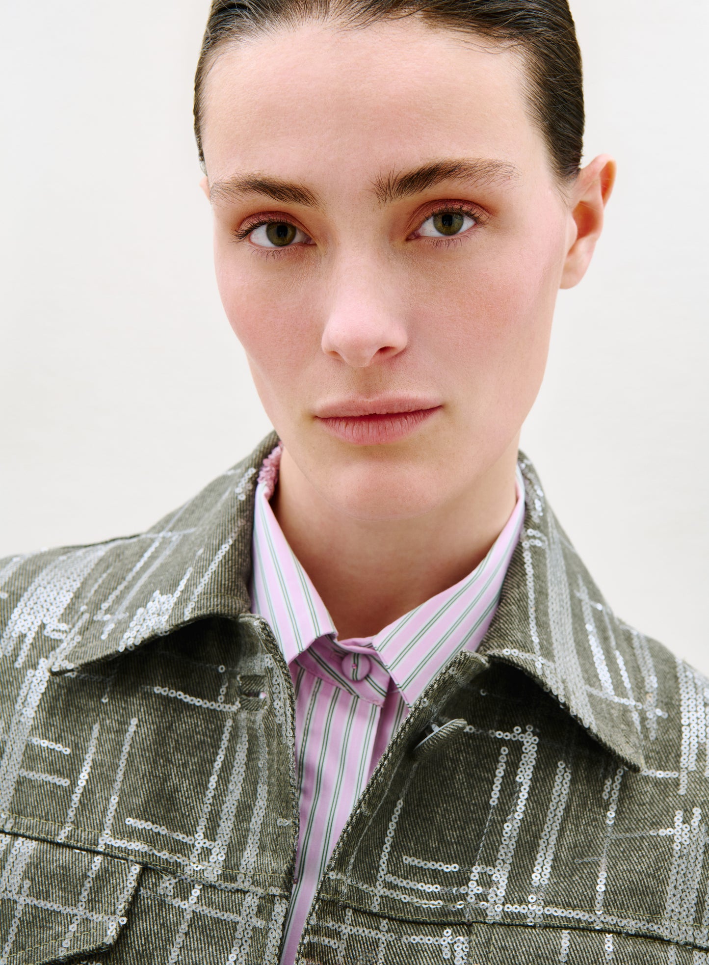 A person with light skin, straight brown hair slicked back, and hazel eyes looks at the camera. They wear the Lesath Jacket—a green plaid piece with a shiny finish—over a pink and white striped collared shirt. The background is plain and light.
