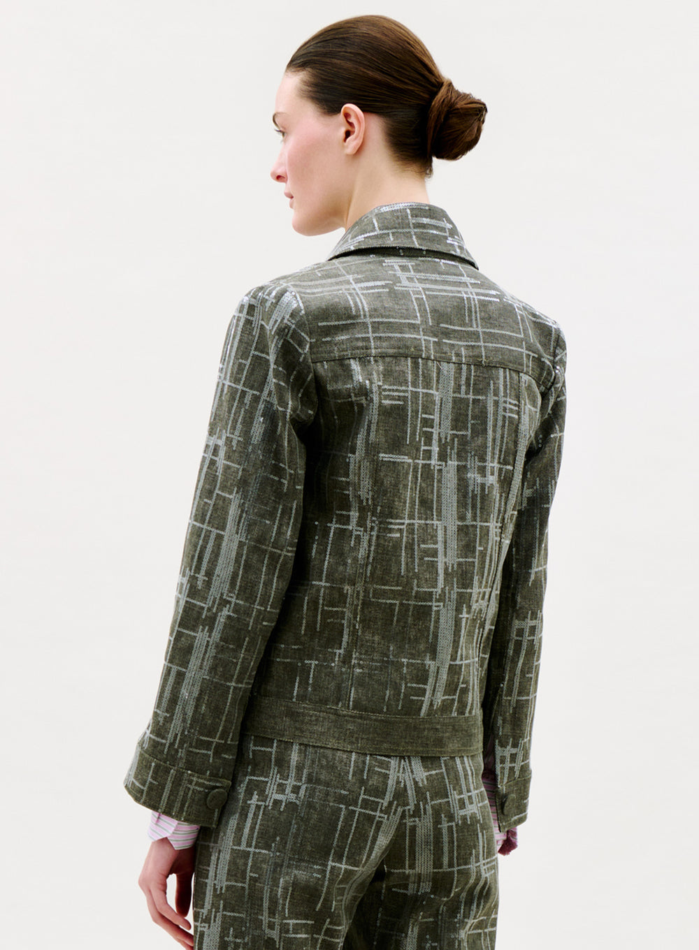A person with fair skin and brown hair in a bun is seen from behind, wearing the Lesath Jacket in gray with matching pants, standing against a plain light background.