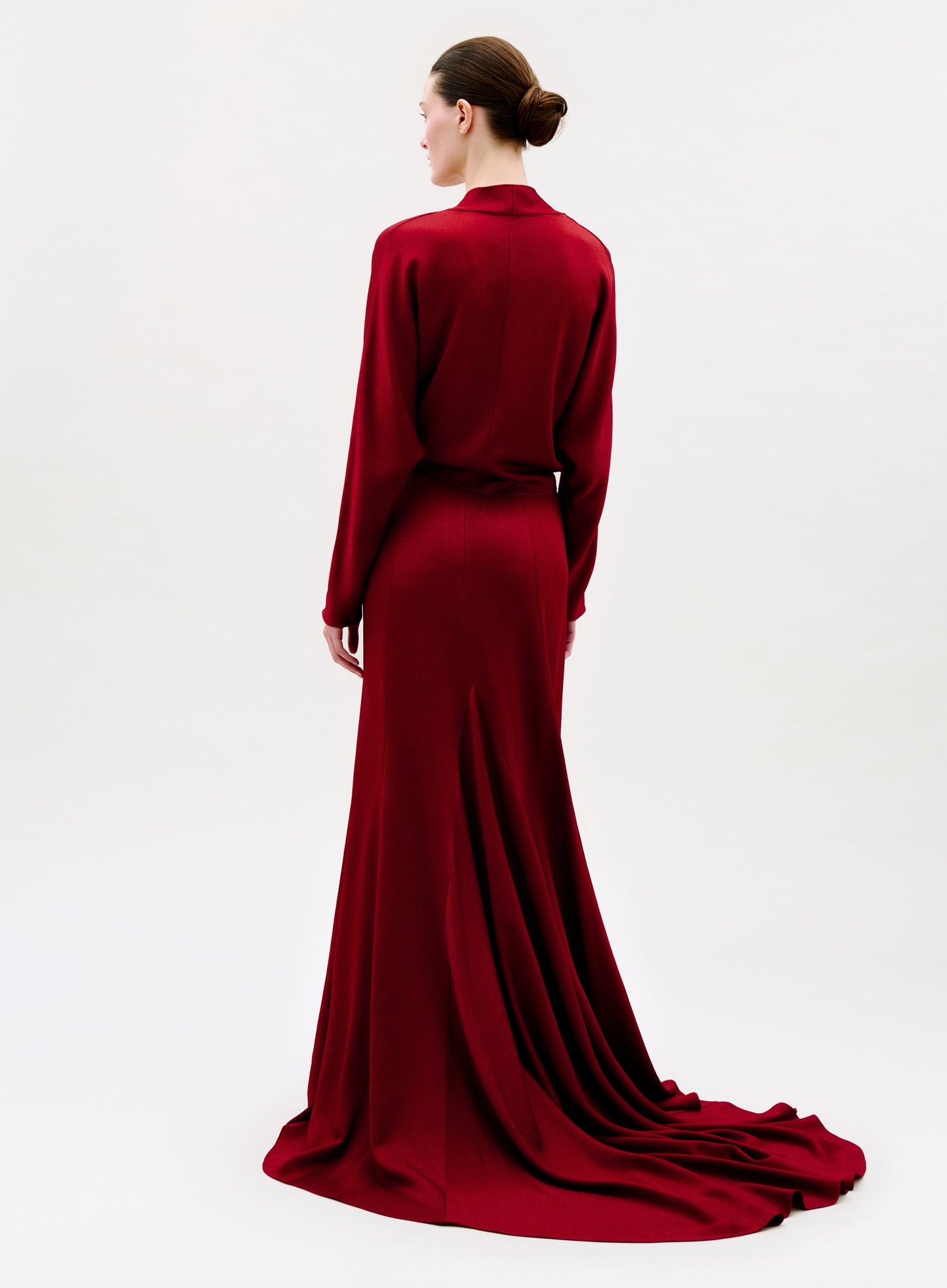 A person with brown hair in a low bun faces away from the camera, standing against a light background and wearing the Marlon Skirt—a long, floor-length deep red skirt made from luxurious Italian fabric.