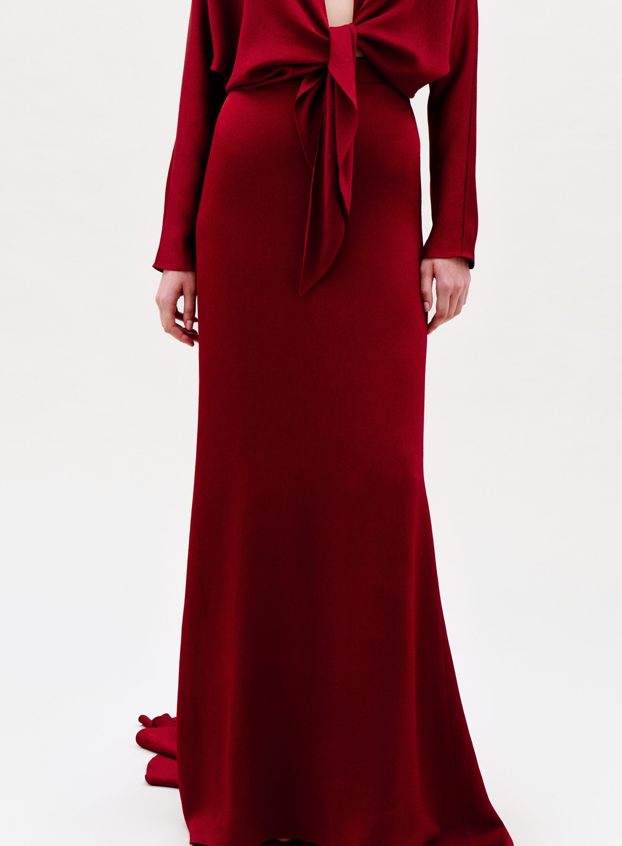 A person models the Marlon Skirt, a long deep red column-style skirt with bow detail, standing against a plain light background. The person's face is not shown.
