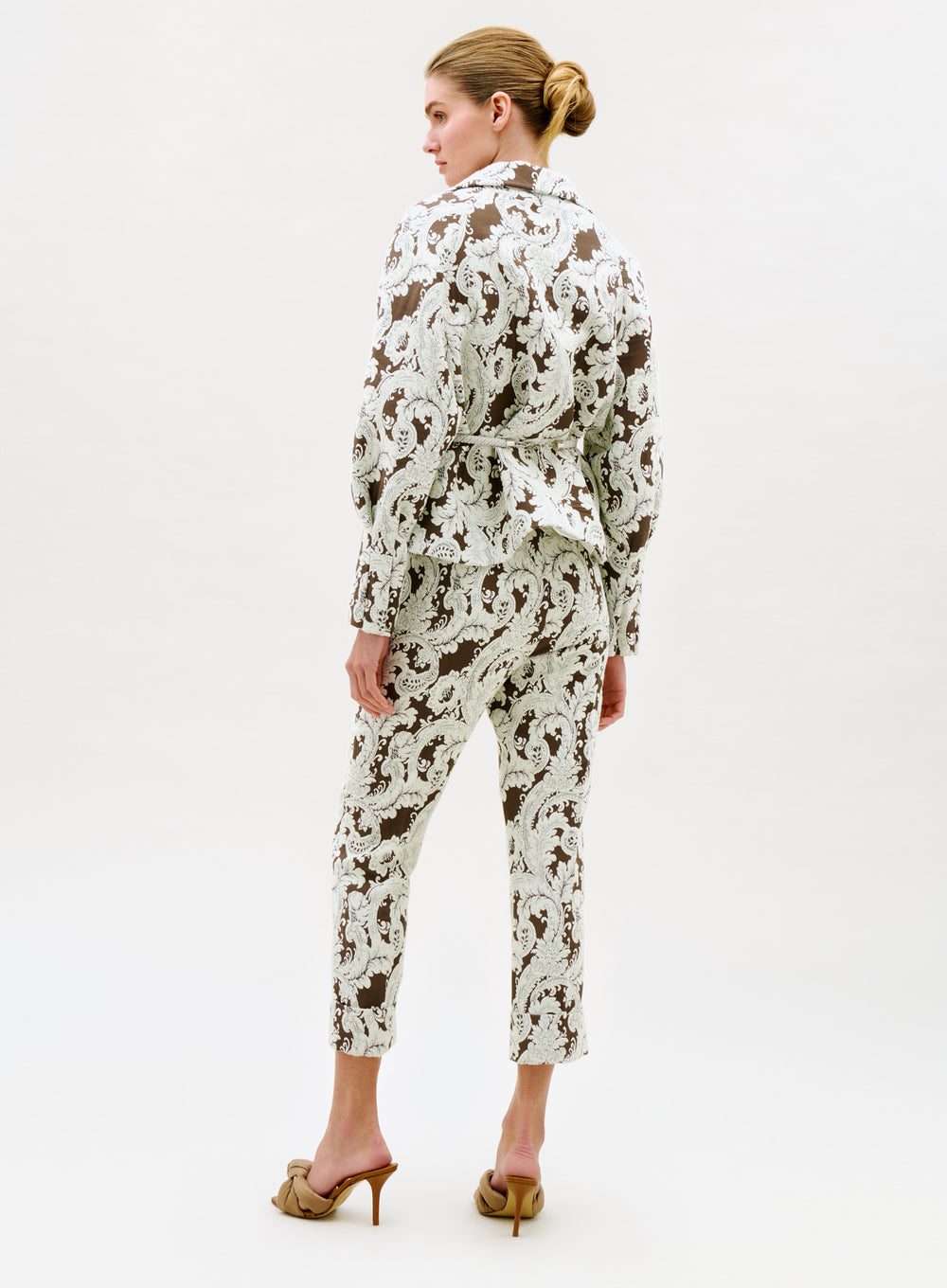 A person stands facing away, wearing the Moad Pant—a white and brown paisley-patterned, tapered suit pant—paired with heeled sandals and a matching jacket. Their hair is in a neat low bun against a plain light background.