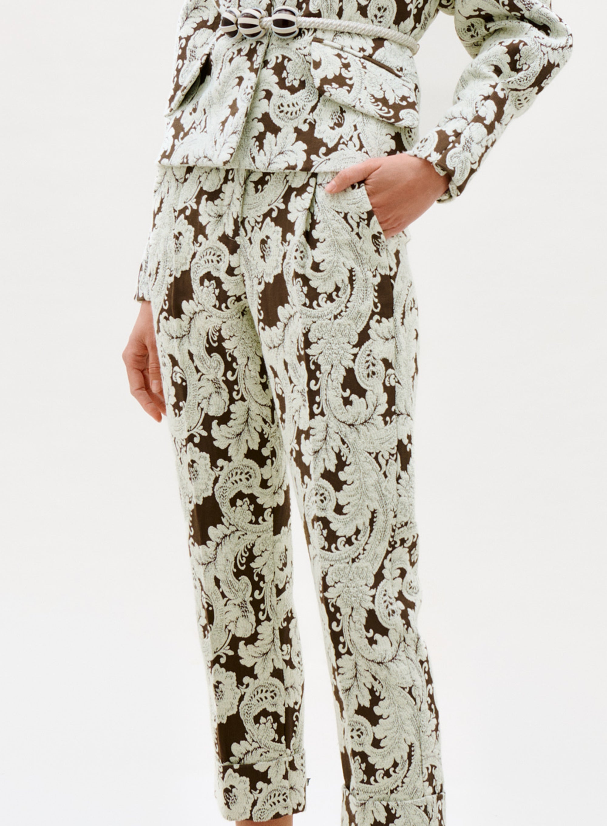 A person wearing the Moad Pant, a matching suit with a light green and brown ornate baroque pattern, featuring fitted trousers and a belted jacket, standing against a plain white background.