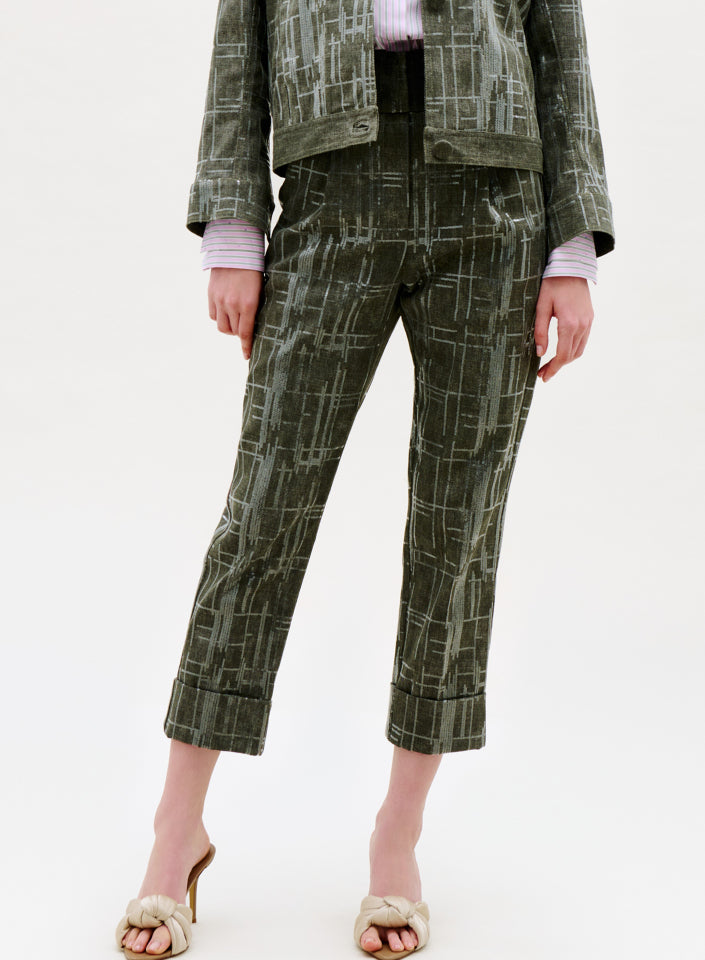 Dressed in the Moad Pant, high waisted cropped olive green pants made from Italian fabric with a white abstract line pattern, paired with a matching jacket, light pink top, and cream heeled sandals, someone stands against a plain background.
