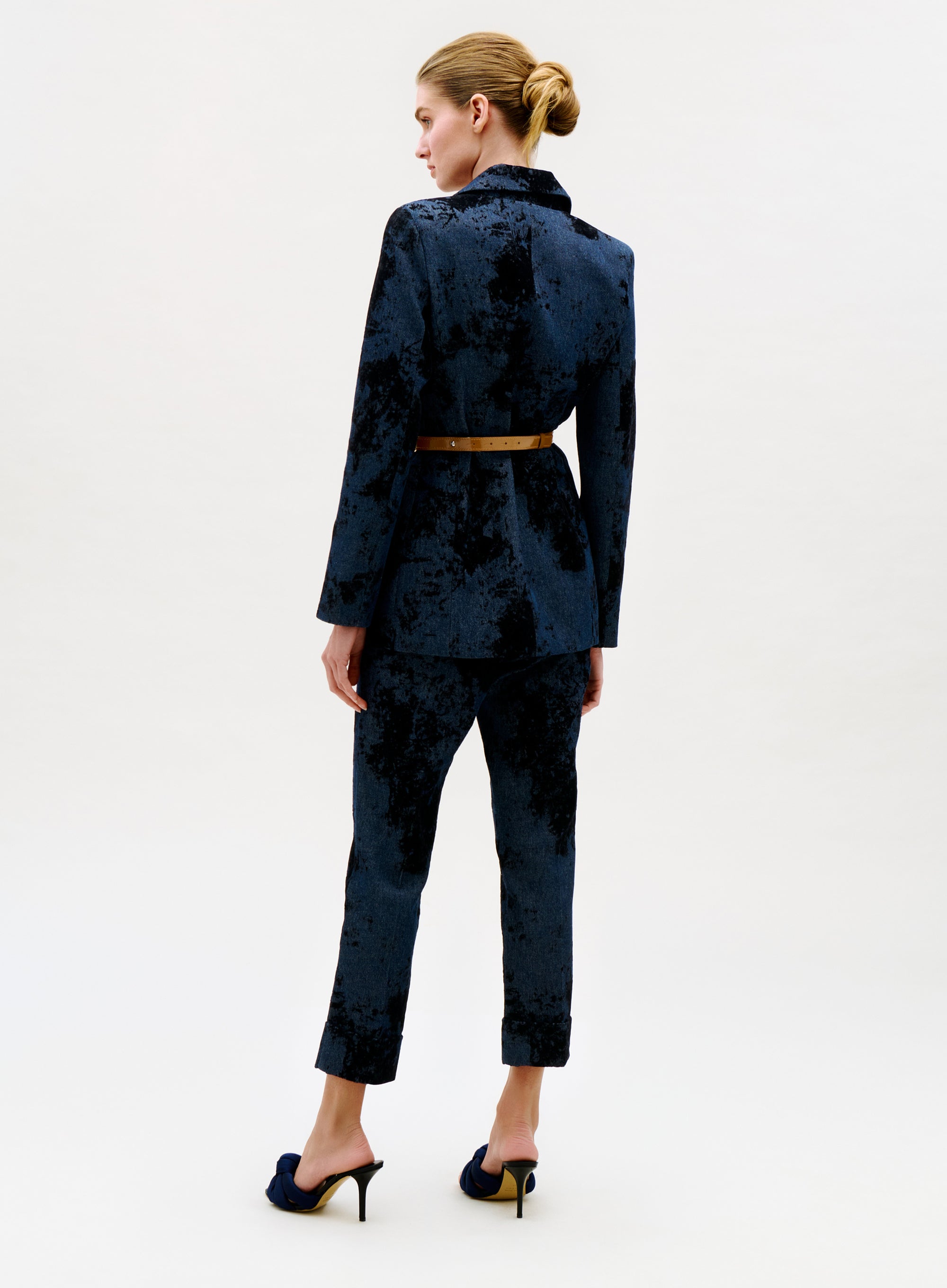 A woman faces away, wearing the Moad Pant—a dark velvet suit with a belted waist and cropped pants. Her blonde hair is in a neat bun, and she pairs black heeled sandals with the outfit against a plain white background.