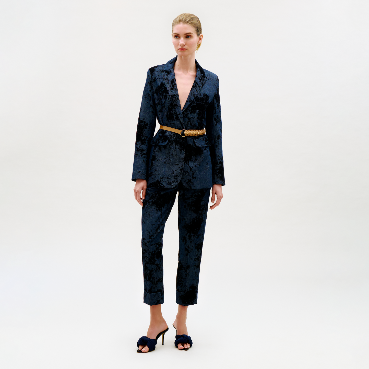 A woman stands against a plain background wearing the Moad Pant, part of a dark velvet pantsuit with a deep V-neck blazer, gold belt, and black high-heeled sandals.