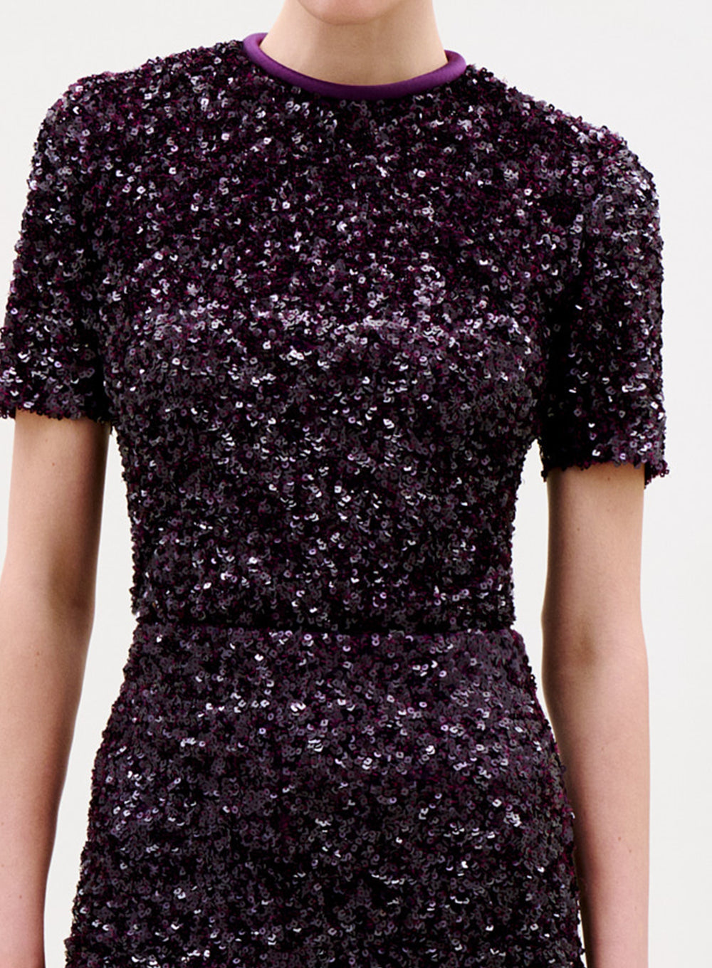 A person wearing the Napoles Blouse, a short-sleeved top with a fitted silhouette and round neckline trimmed in light purple. The dark purple, densely shimmering sequins give this elegant blouse a chic look.