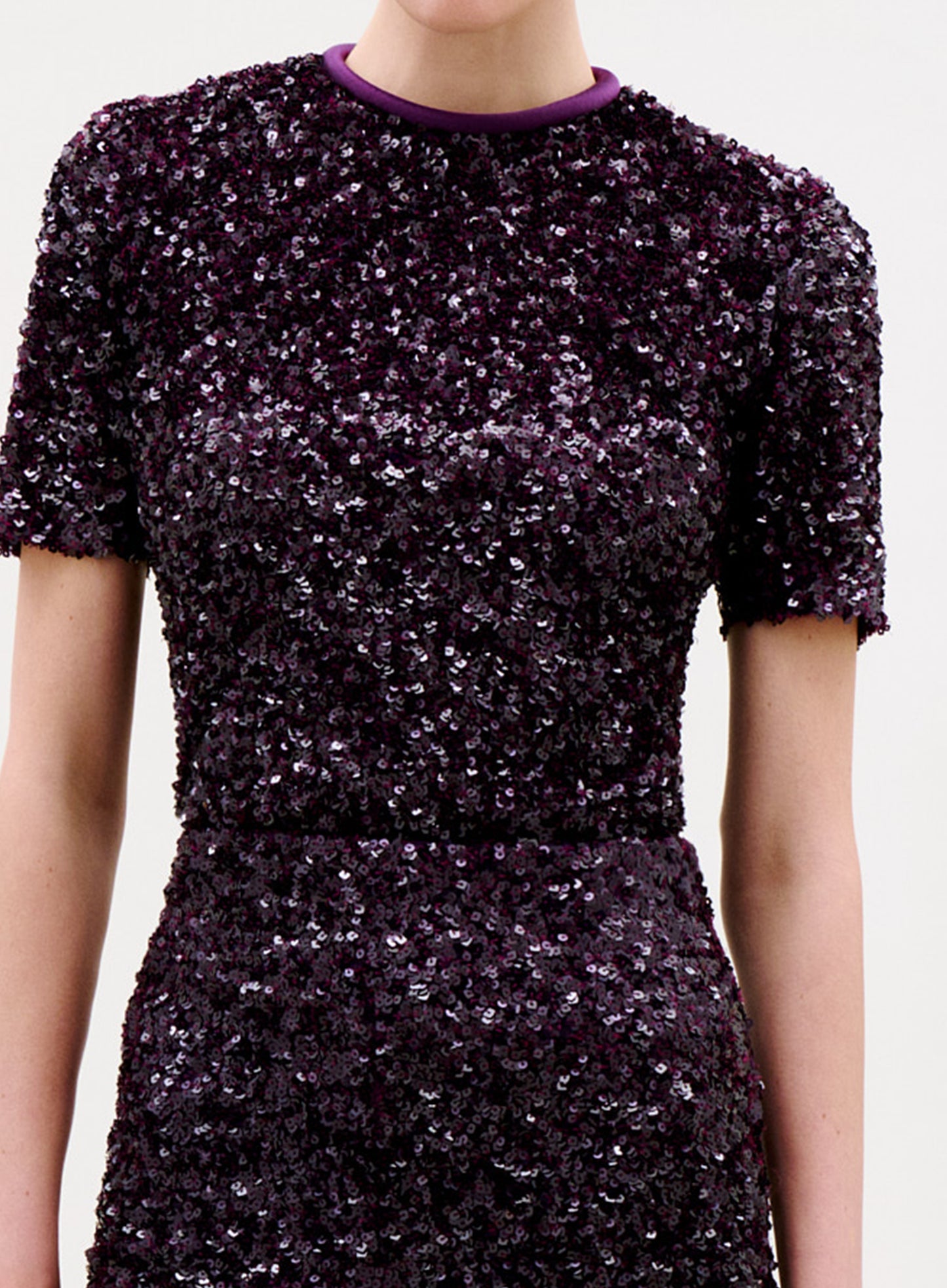 A person wearing the Napoles Blouse, a short-sleeved top with a fitted silhouette and round neckline trimmed in light purple. The dark purple, densely shimmering sequins give this elegant blouse a chic look.