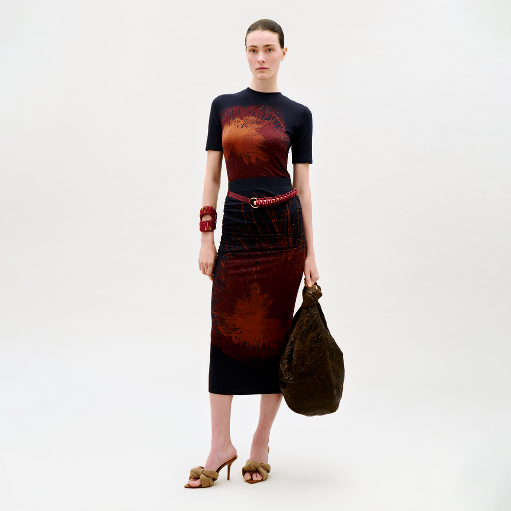 A woman stands against a plain background, wearing the Niove Skirt in a dark hue with red abstract patterns, brown heeled sandals, a chunky red bracelet, a red chain belt, and holding a large textured brown bag.