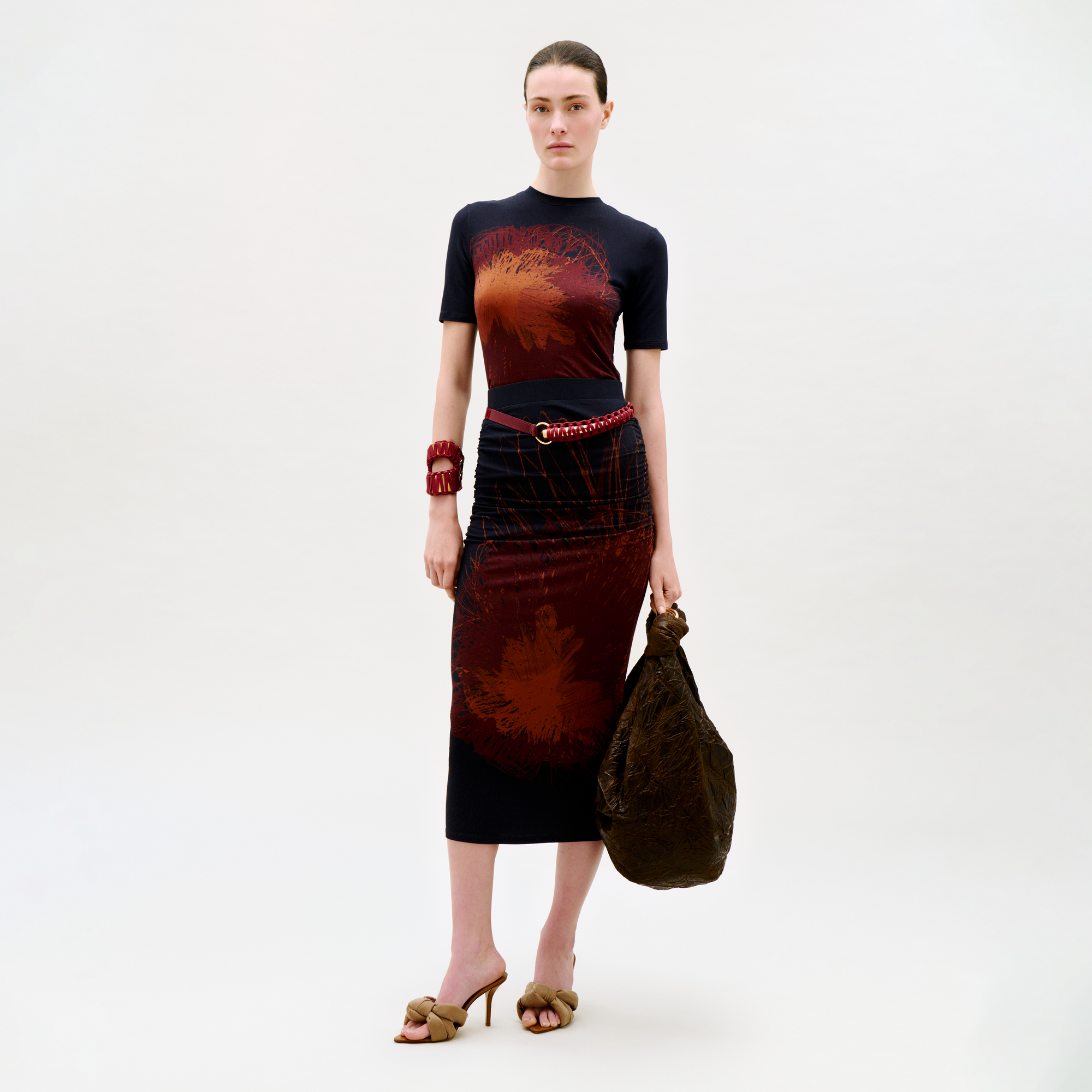 A woman stands against a plain background, wearing the Niove Skirt in a dark hue with red abstract patterns, brown heeled sandals, a chunky red bracelet, a red chain belt, and holding a large textured brown bag.