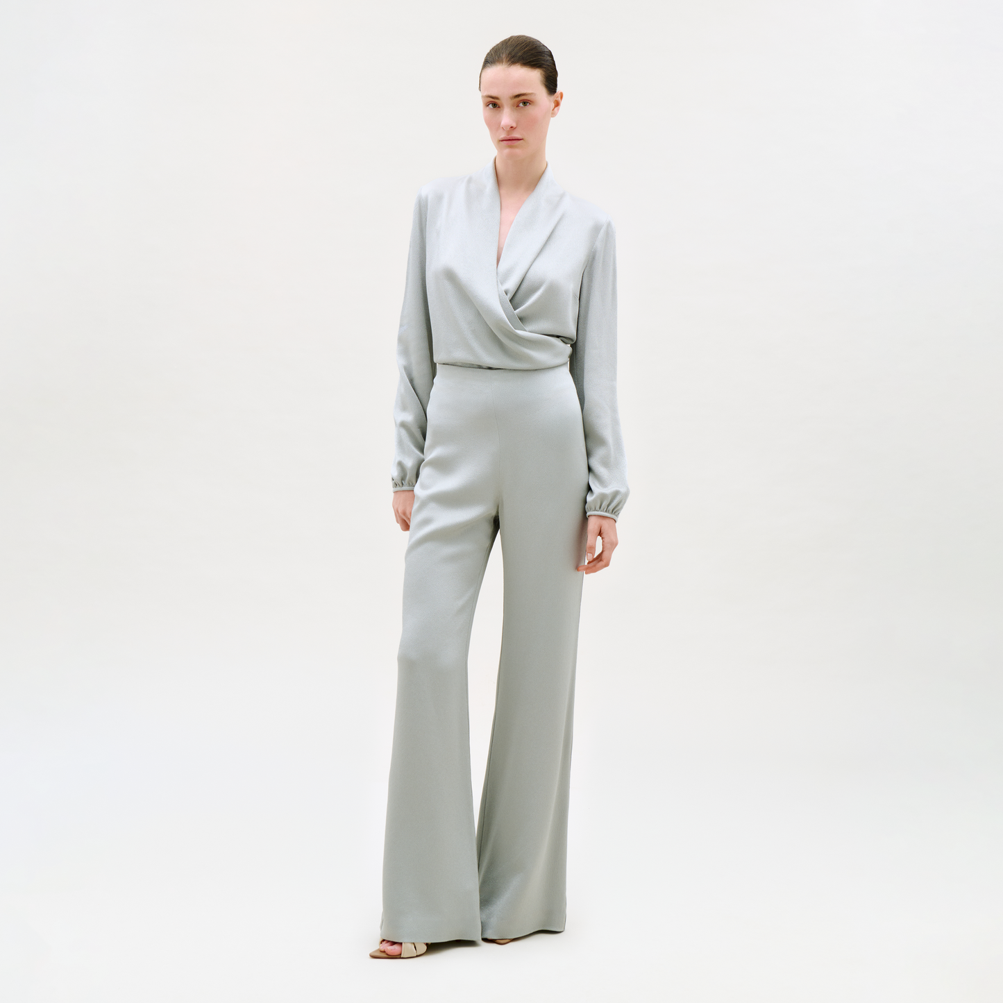 A woman with her hair pulled back and a neutral expression stands against a plain light background, wearing the Palermo Pant—light gray, wide-leg style—paired with a matching long-sleeve wrap-front top and open-toe heels.