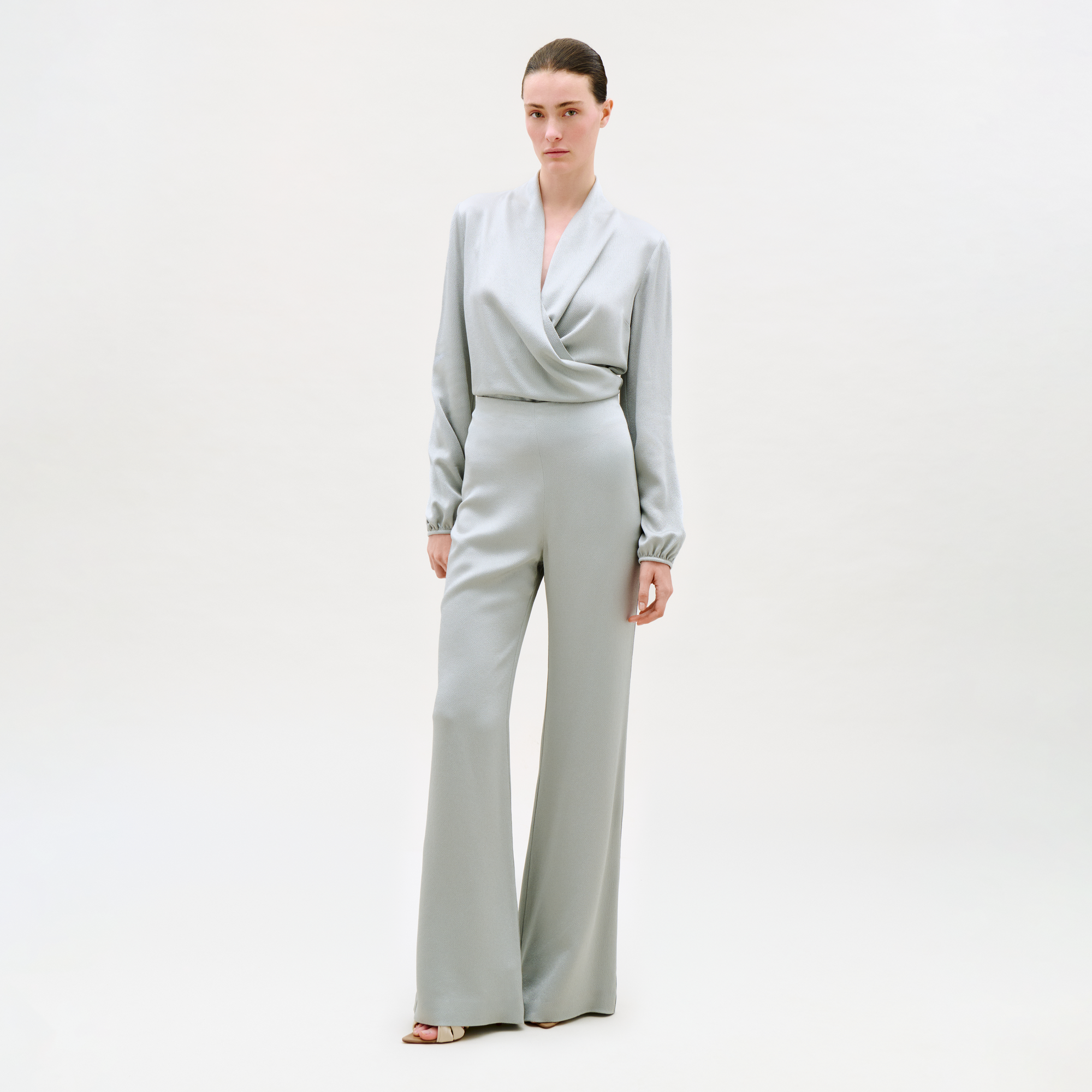A woman with her hair pulled back and a neutral expression stands against a plain light background, wearing the Palermo Pant—light gray, wide-leg style—paired with a matching long-sleeve wrap-front top and open-toe heels.