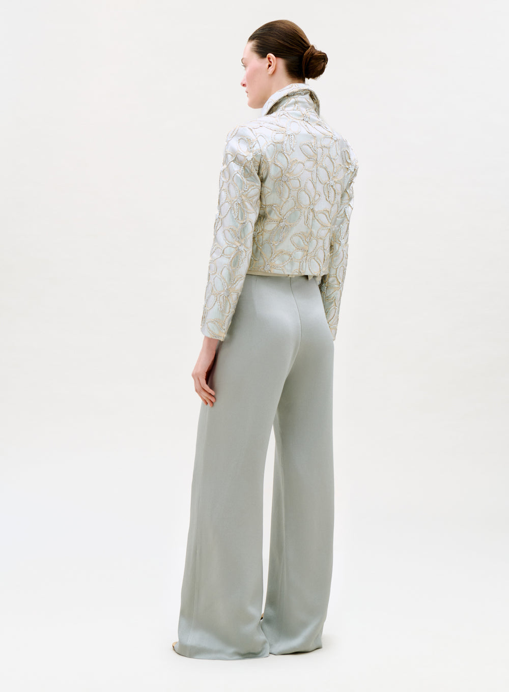 A woman with a neat low bun stands facing away, wearing the Palermo Pant—high-waisted, wide-leg pants made from exquisite Italian fabric—paired with a light embroidered jacket against a plain background.