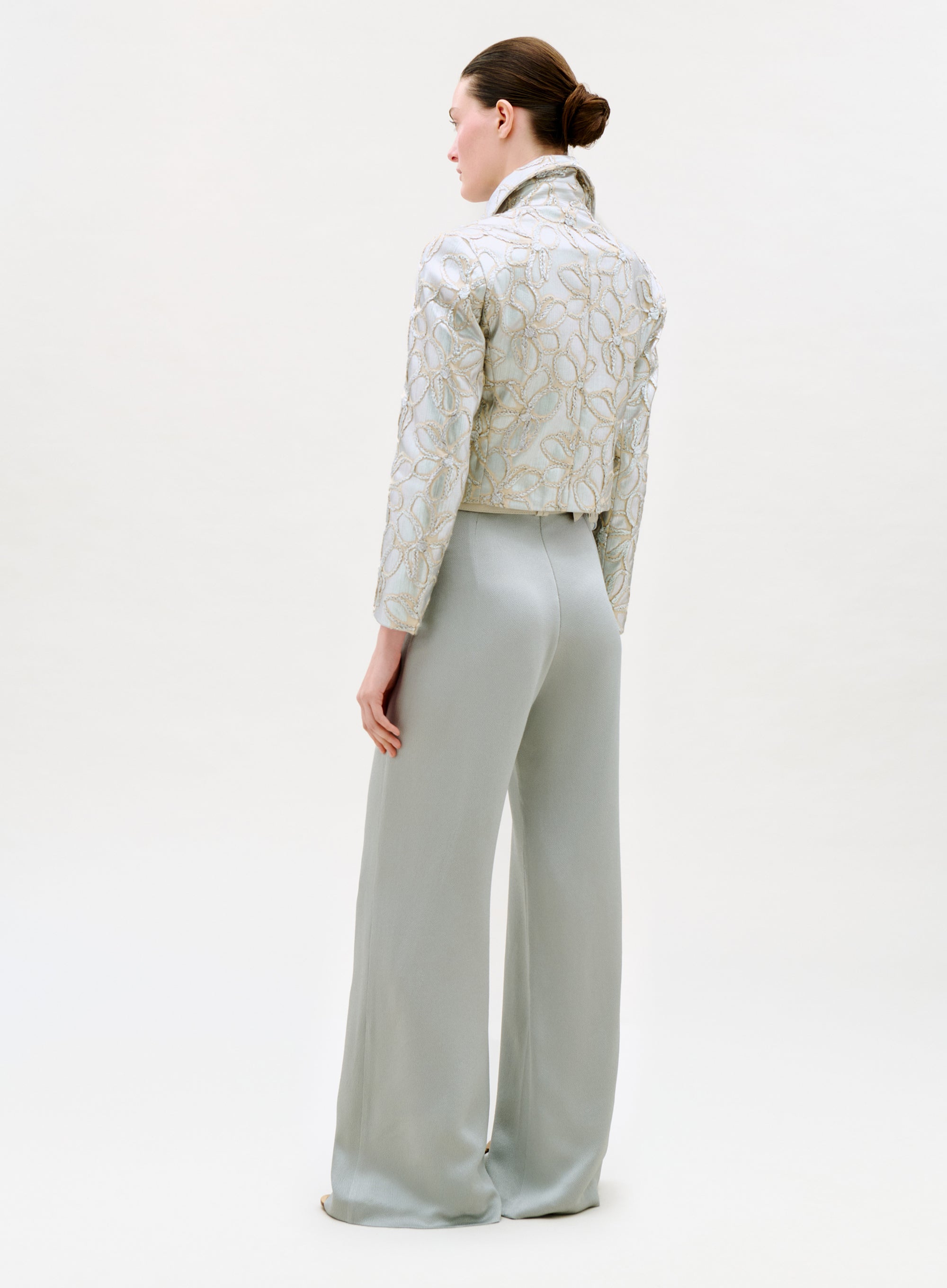 A woman with a neat low bun stands facing away, wearing the Palermo Pant—high-waisted, wide-leg pants made from exquisite Italian fabric—paired with a light embroidered jacket against a plain background.