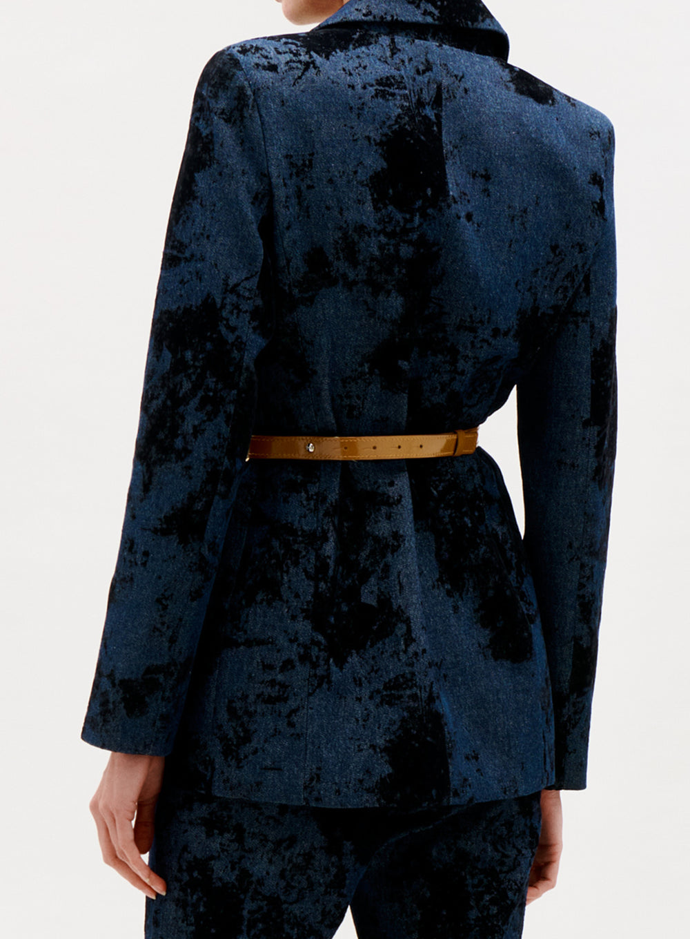 A person is shown from the back wearing the Rebeca Jacket, a blue and black patterned velvet blazer with matching pants and a tan belt, against a plain light background.