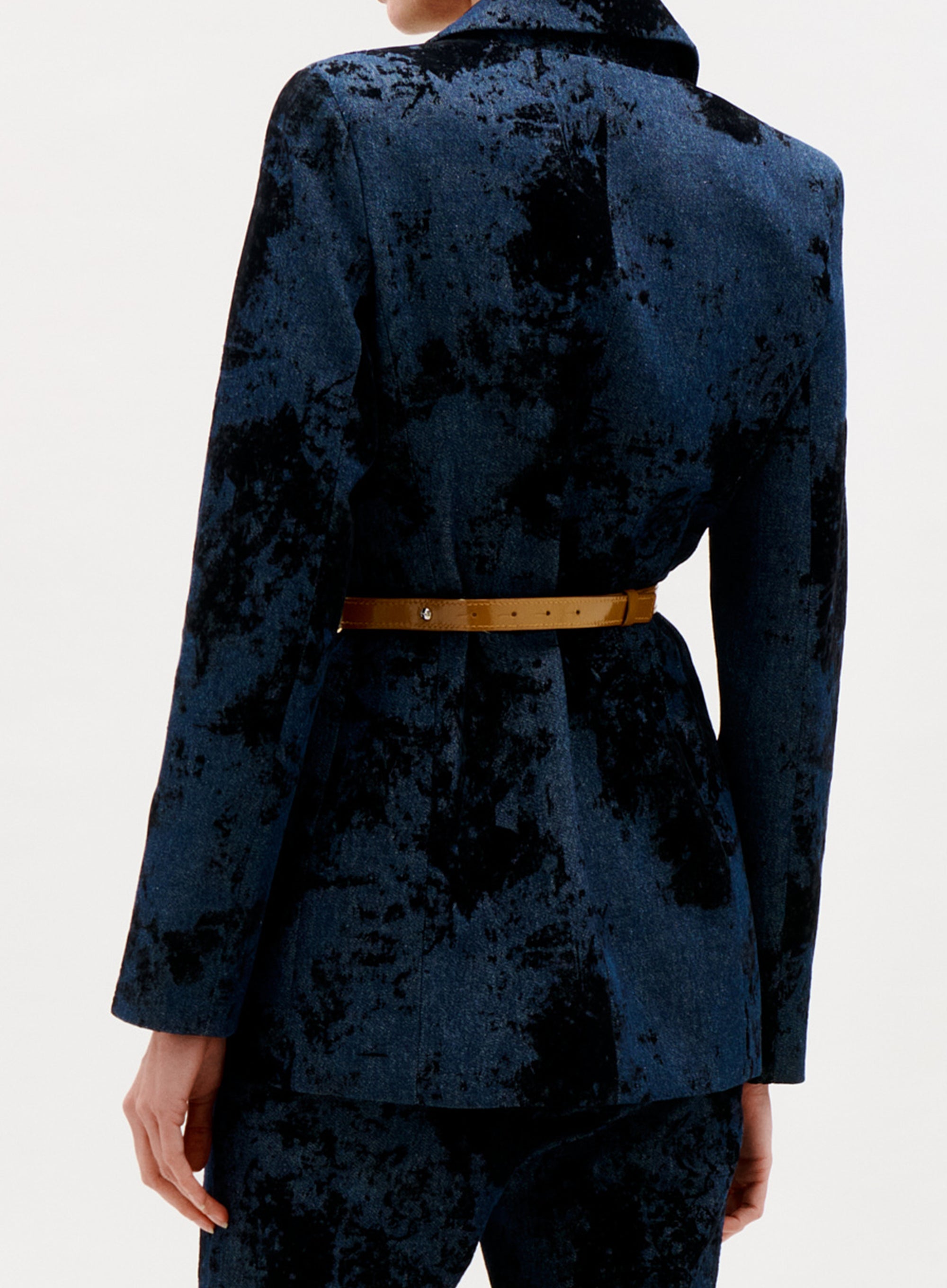 A person is shown from the back wearing the Rebeca Jacket, a blue and black patterned velvet blazer with matching pants and a tan belt, against a plain light background.