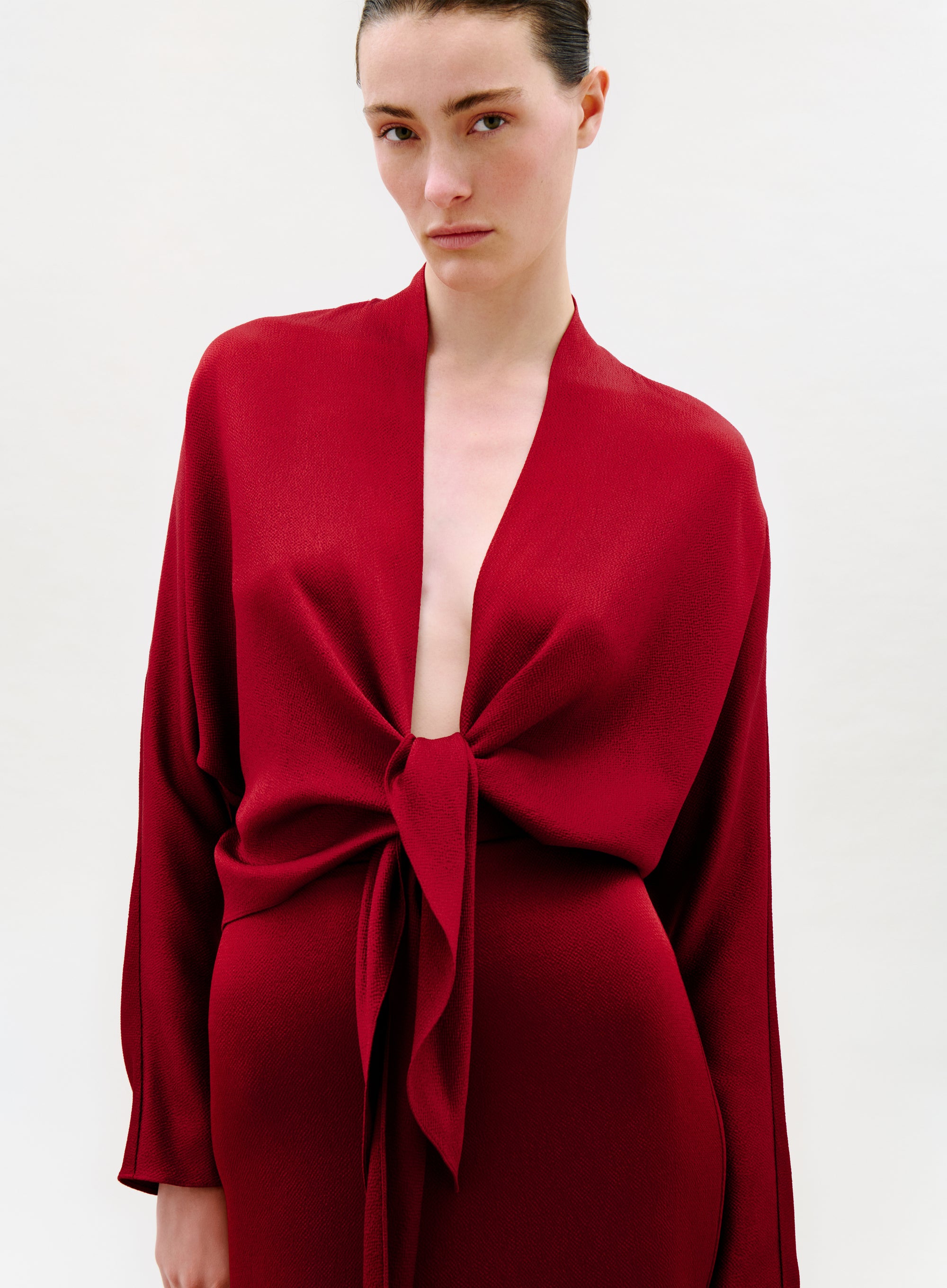 A woman with dark hair pulled back wears the deep red Reiko Blouse featuring long sleeves, an open front, and a large knotted waist tie, standing against a plain white background with a neutral expression.