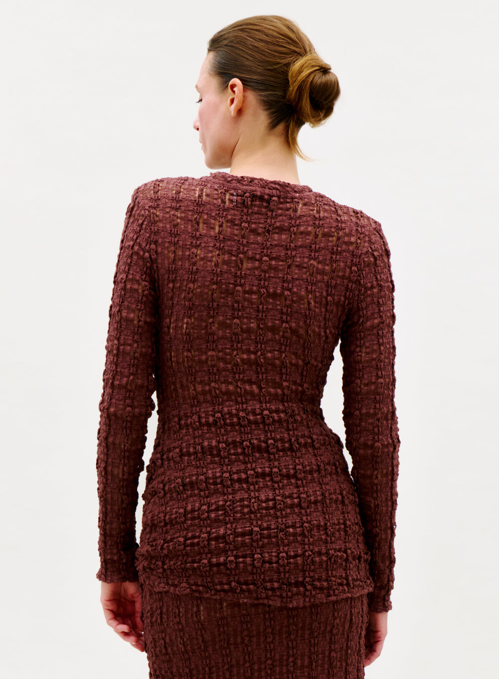 A woman with brown hair in a neat bun is seen from behind, wearing the Roma T-Shirt—a textured, long-sleeve top made in Colombia—against a plain white background.