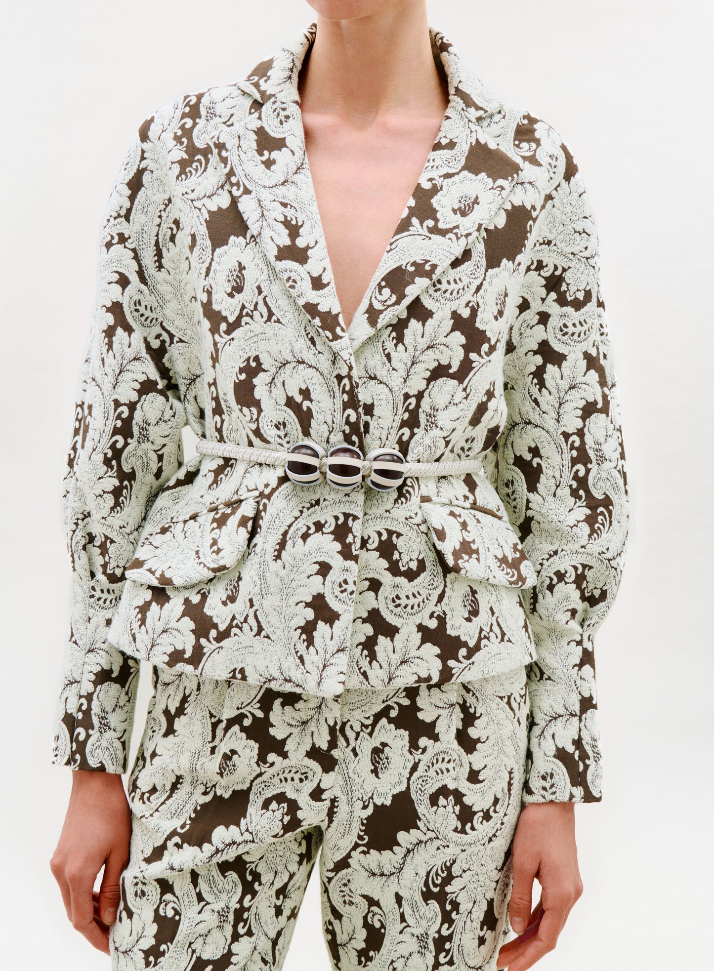 A person wearing the Noureen Jacket, featuring a brown hue with white ornate floral patterns, crafted from luxurious Italian fabric and complemented by a matching belt with a decorative silver buckle, stands against a plain background.
