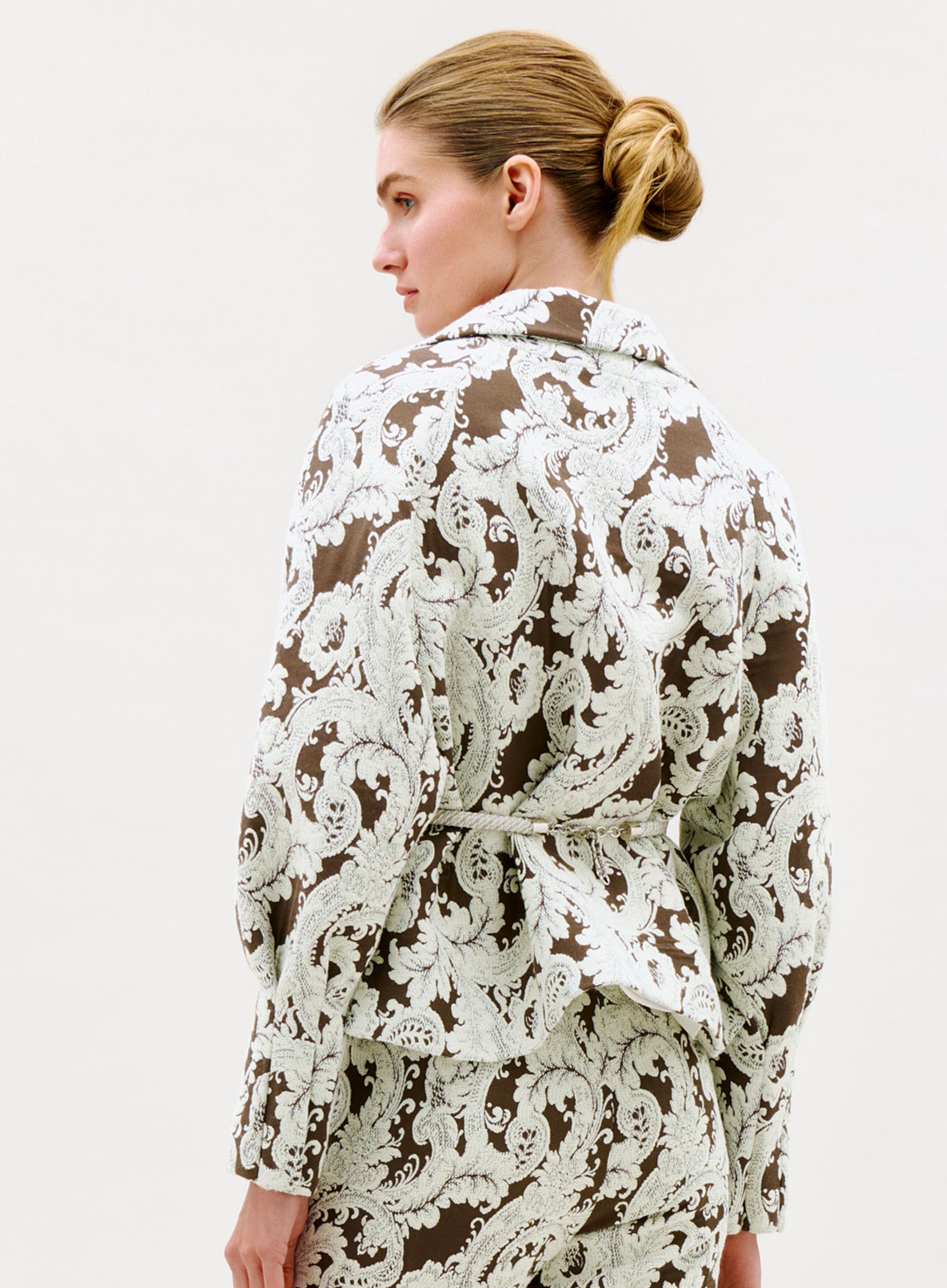 A woman with blonde hair in a low bun is seen from behind and slightly to the side, wearing the Noureen Jacket—a brown and white paisley-patterned suit jacket with a belt—set against a plain light background.