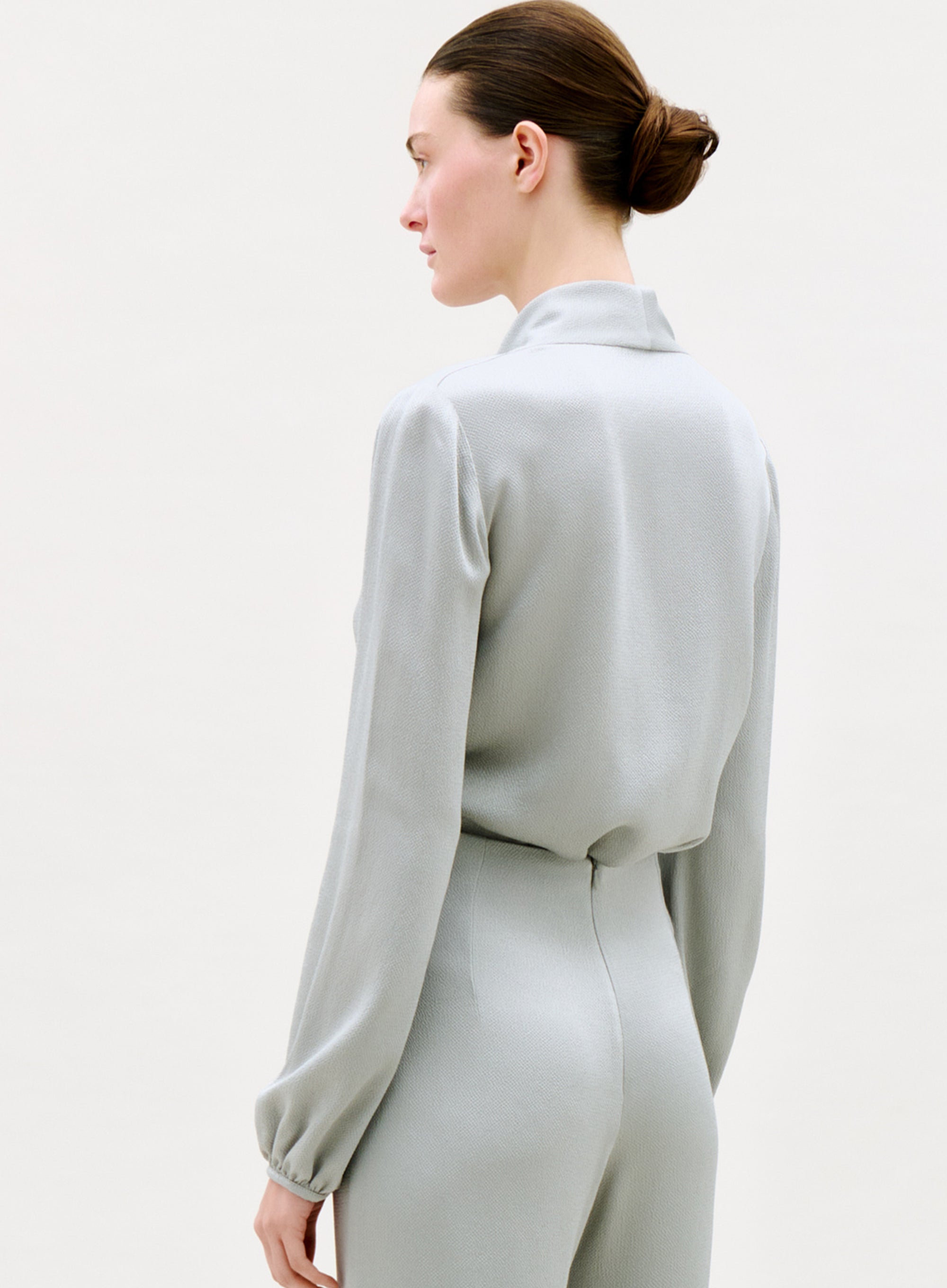 A person with brown hair in a low bun wears the light gray, long-sleeved Tea Blouse and matching high-waisted pants, shown from the back and side against a plain light background.