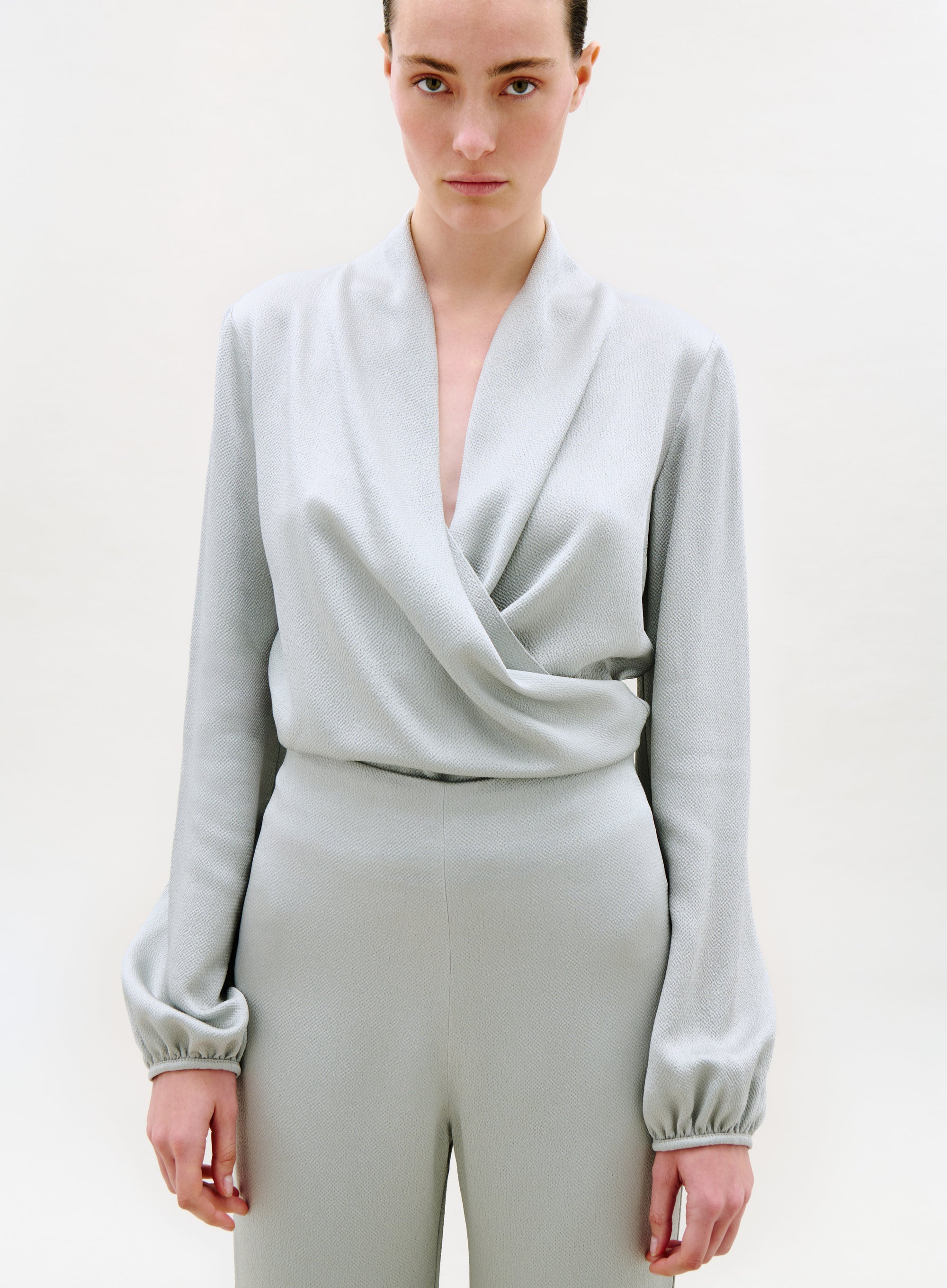 A person with a neutral expression stands against a plain light background, wearing the light gray, long-sleeved Tea Blouse with a deep V-neck and matching high-waisted pants. Their arms are relaxed by their sides.